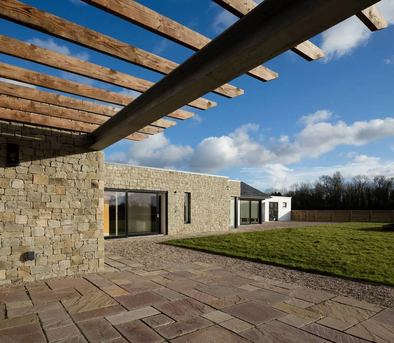 Castleknock, Dublin 15 — A modern house with stone and white exterior walls, large glass doors, a wooden pergola, a stone patio, green grass, and a clear blue sky with scattered clouds.