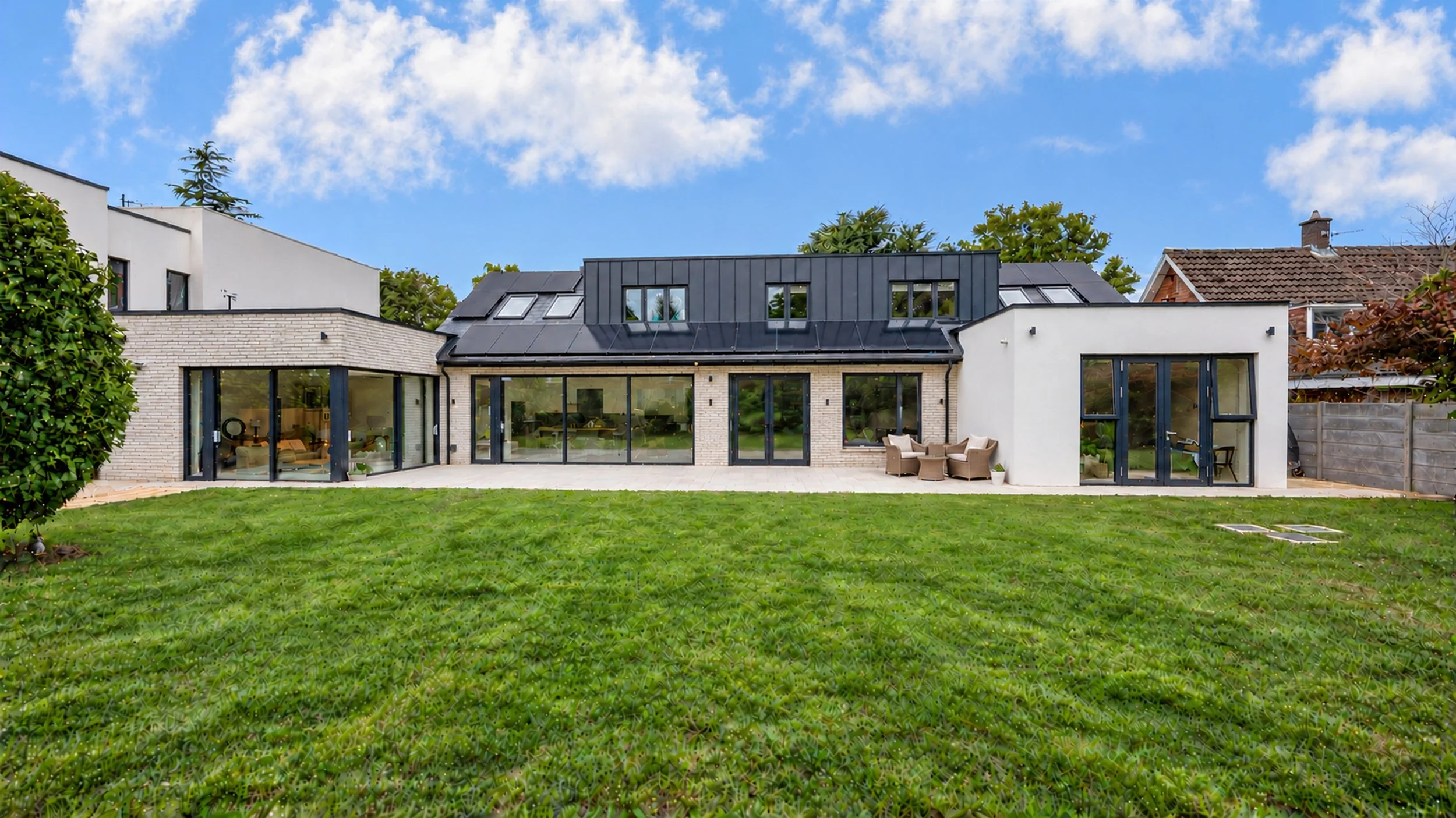 Cherrymount — Modern house with large glass doors, a grassy backyard, patio furniture, and neighboring houses, under a blue sky with clouds.