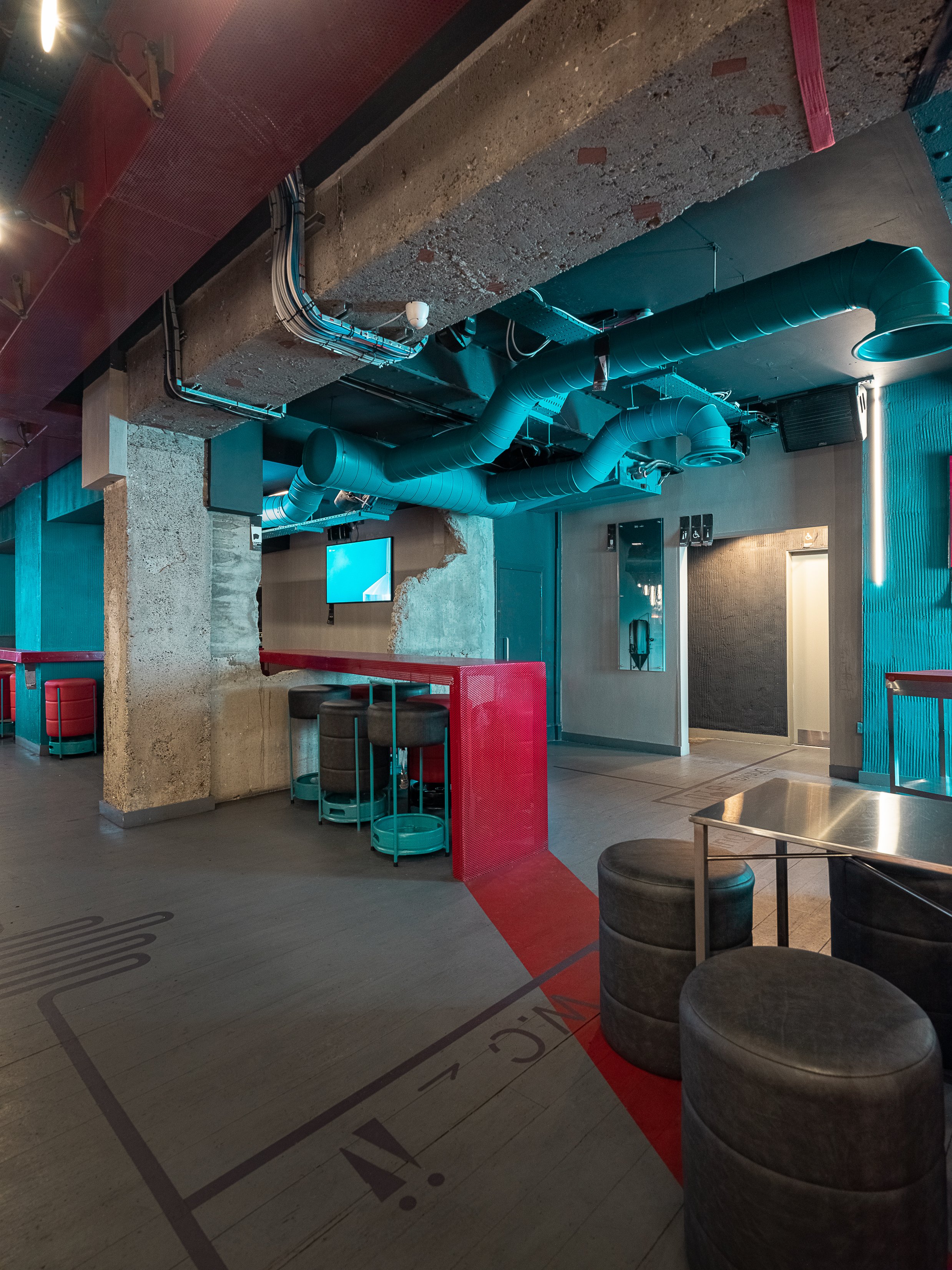 Tapped, Grafton Street, Dublin — Modern interior space with exposed concrete pillar, green ventilation ducts on ceiling, red partition, computer monitors, black stools, and gray cushioned seating.