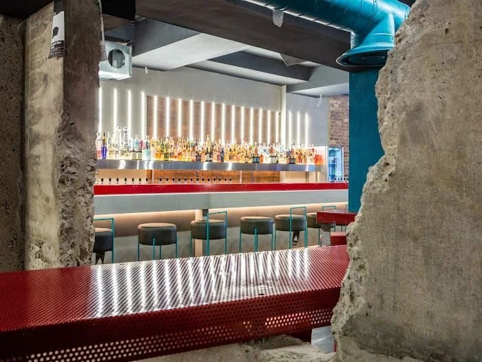 Tapped, Grafton Street, Dublin — Bar with illuminated bottle shelves, barstools and a red perforated metal counter viewed through a restored original brick archway.
