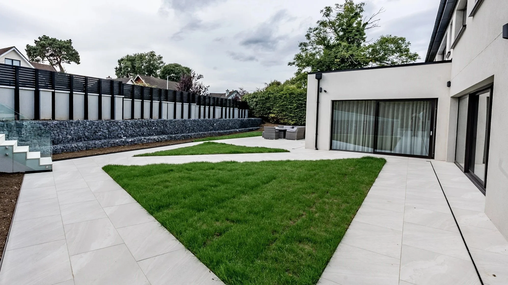Salzburg, Dundrum — Modern house backyard with white tile patio, green lawn, outdoor seating area, and tall black and white fences under cloudy sky.
