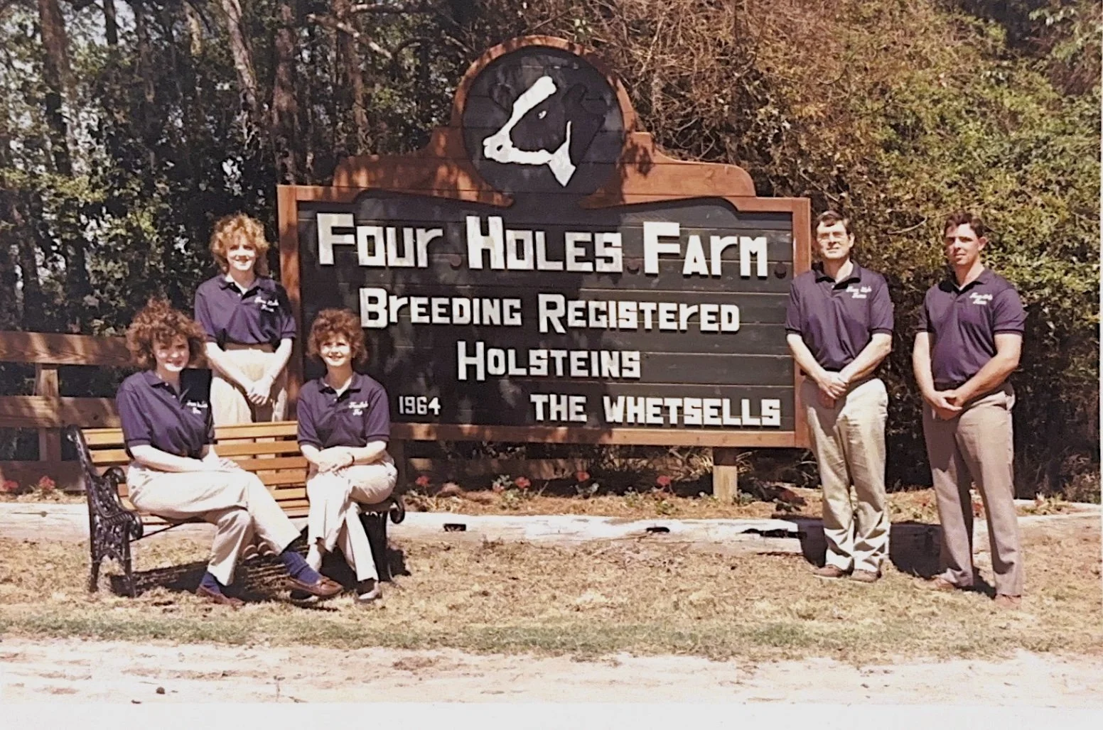 Group of five people wearing matching purple shirts standing and sitting next to a large wooden sign that reads "Four Hollows Farm Breeding Registered Holsteins The Whetstolls 1964"; a black and white logo of a cow's head at the top of the sign; trees in the background.