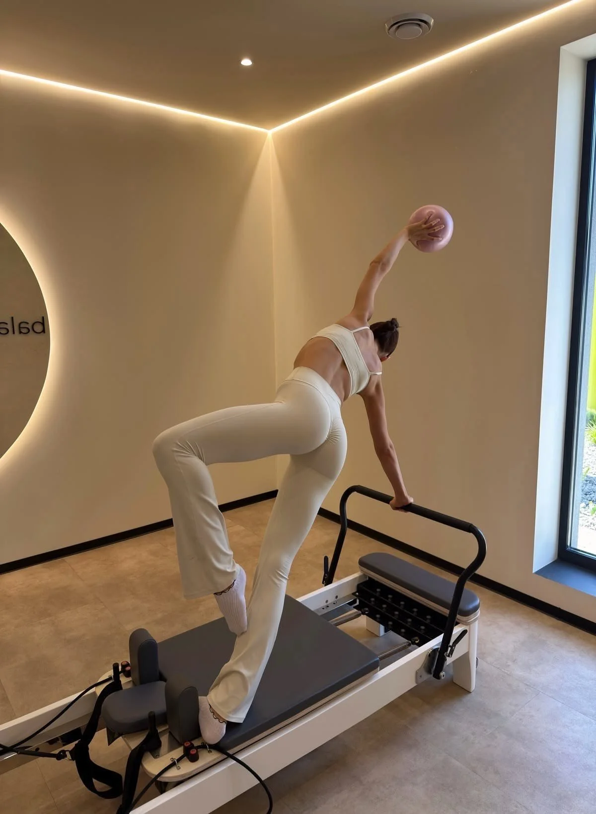 Vrouw die een oefening doet op een Pilates reformer in een fitnessruimte, met een roze bal in haar hand en een grote venster met uitzicht op de buitenkant.