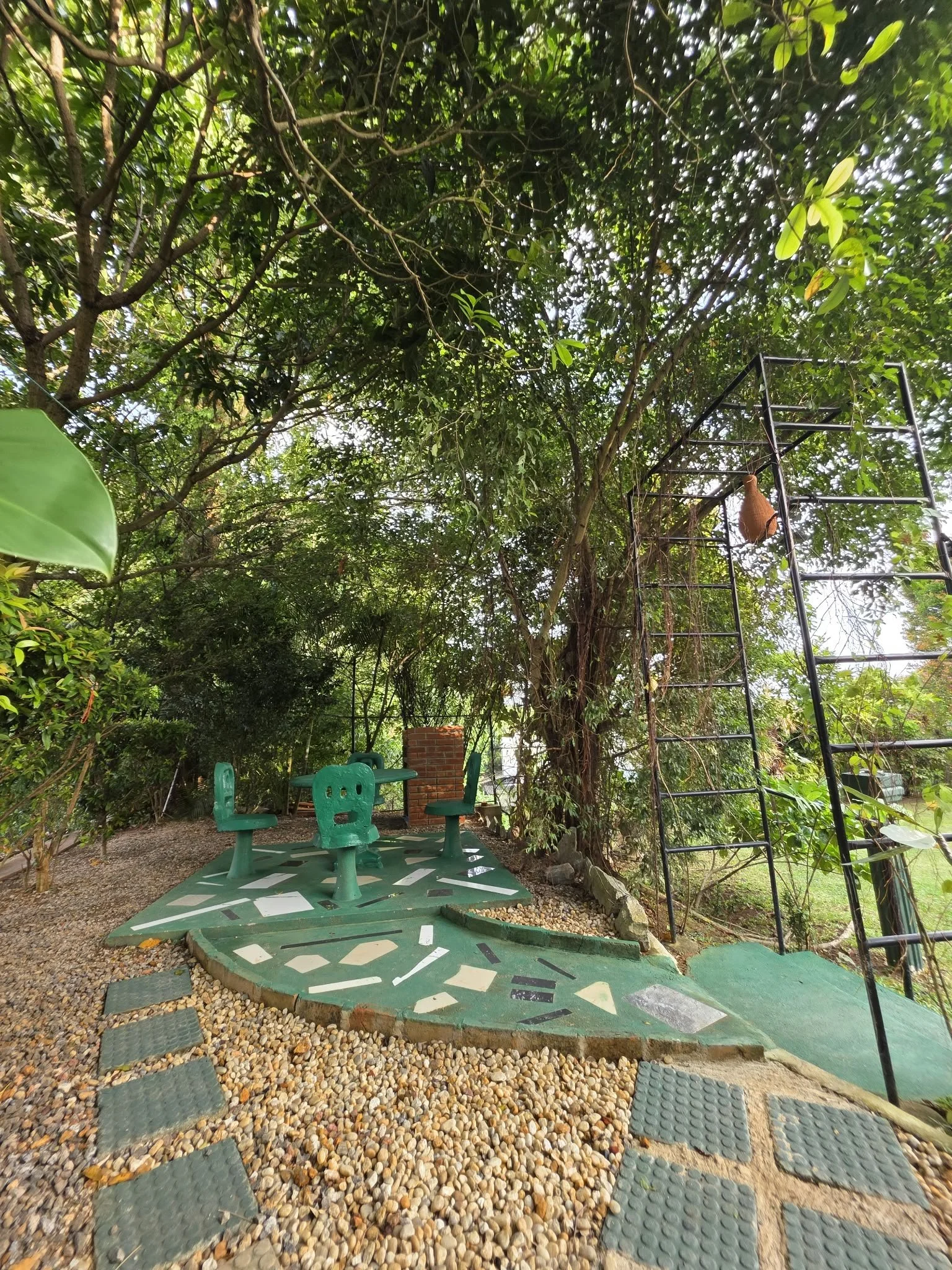 Outdoor park area with four green chairs arranged in a circle, a gravel pathway with stepping stones, a tall metal ladder, trees with dense green foliage, and a small brick structure, all surrounded by lush greenery.