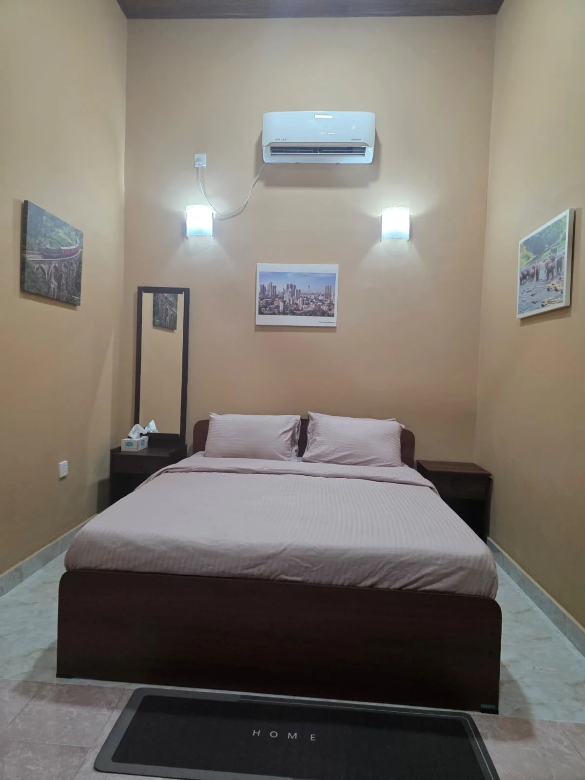 A bedroom with a bed, two pillows, nightstands on each side, a large mirror, and wall art including three framed pictures. An air conditioner is mounted on the wall above the bed, with lights on either side.