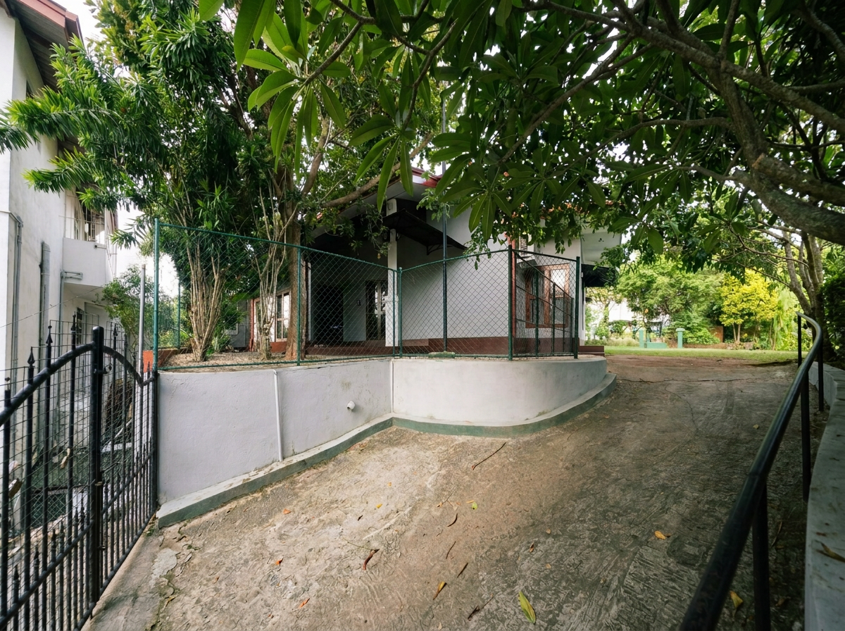 A residential house with trees and greenery, surrounded by a metal fence, and a sloped concrete pathway leading to the entrance.