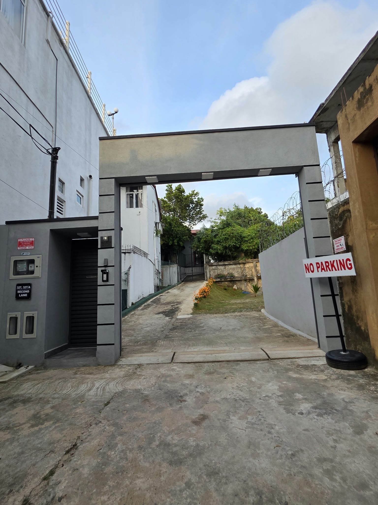 A parking lot with a concrete ground, a gate archway, and a small garden with orange flowers. There are signs that say 'No Parking' near the gate, and a building on the left with utility boxes and pipes.