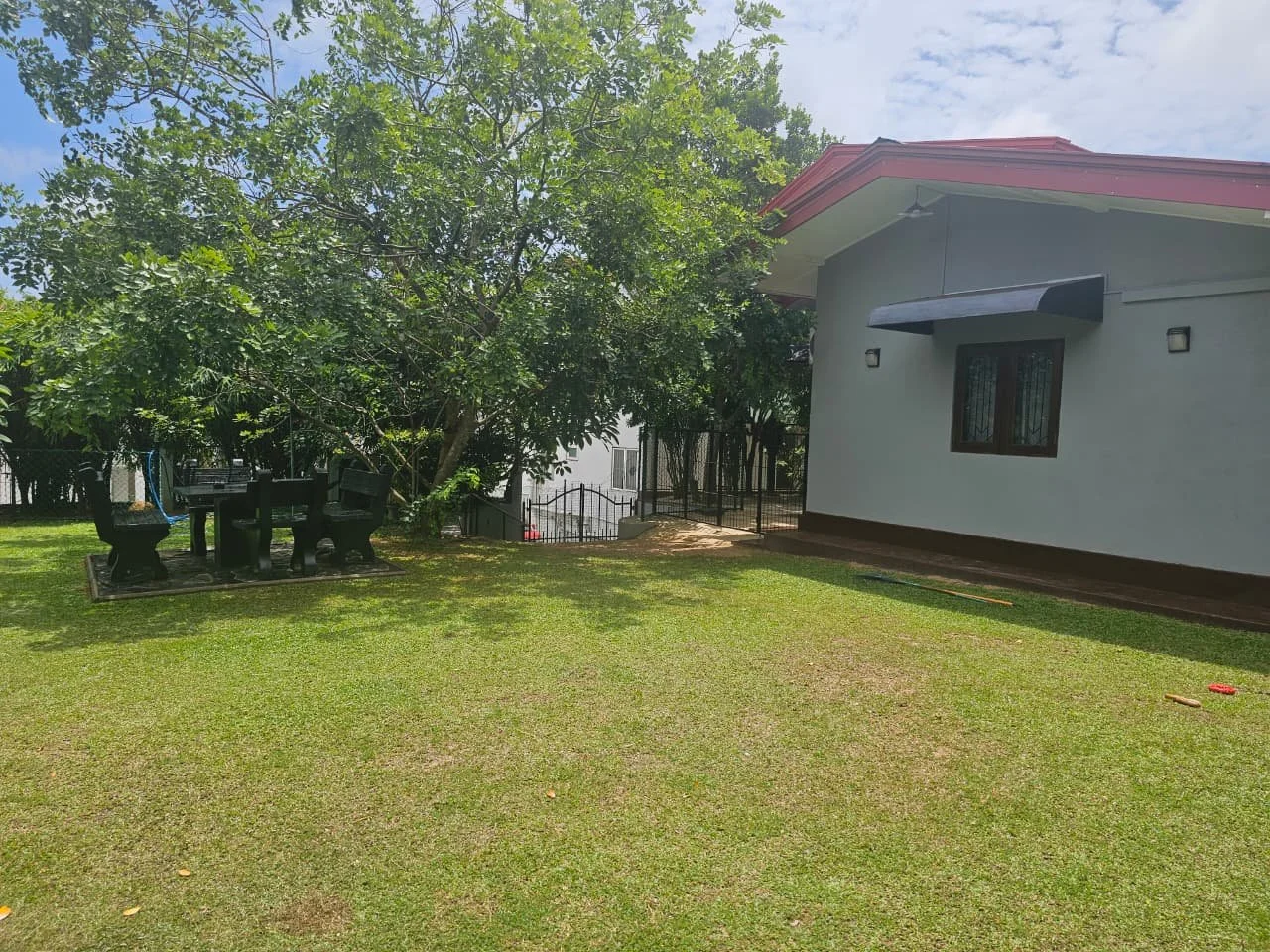 A backyard with green grass, a large tree, an outdoor dining table with chairs, and a house with white walls and a red roof under a partly cloudy sky.