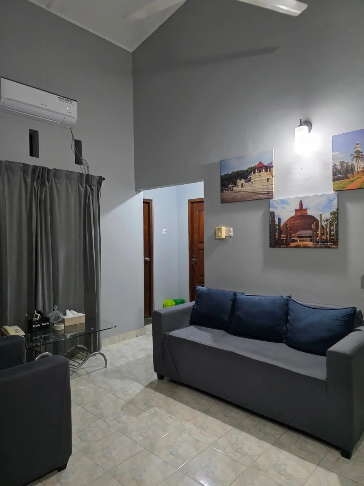 Living room with gray walls, a gray sofa with blue cushions, a glass coffee table, wall art depicting landmarks, dark curtains, and wooden doors.