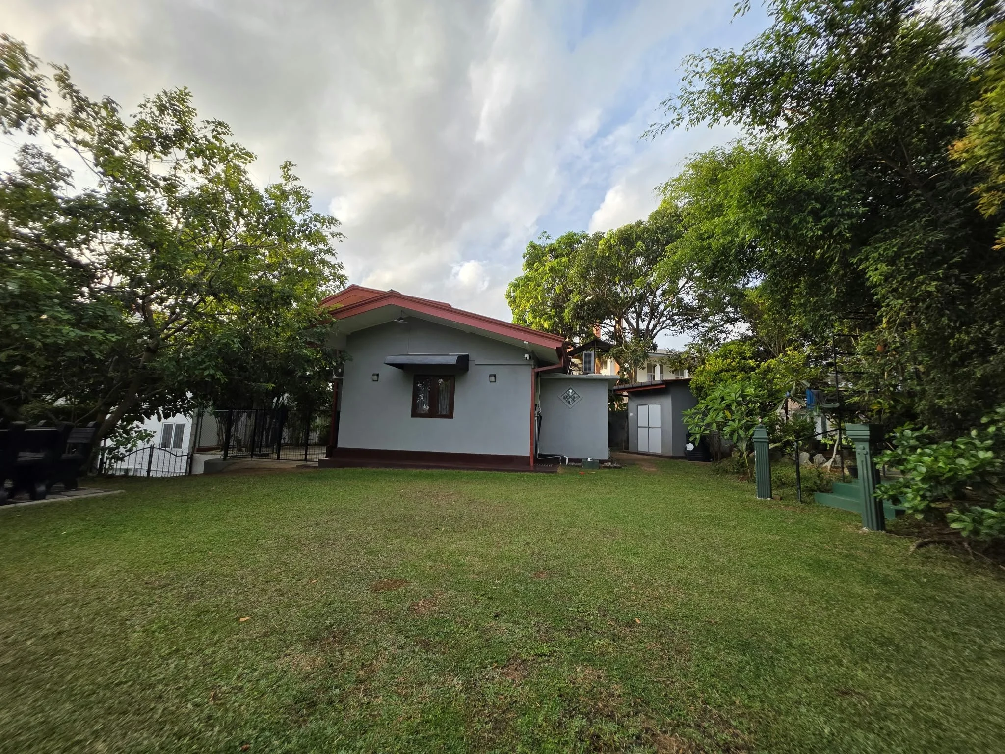 A backyard with a lawn, trees, a small gray house with a red roof, and a cloudy sky.