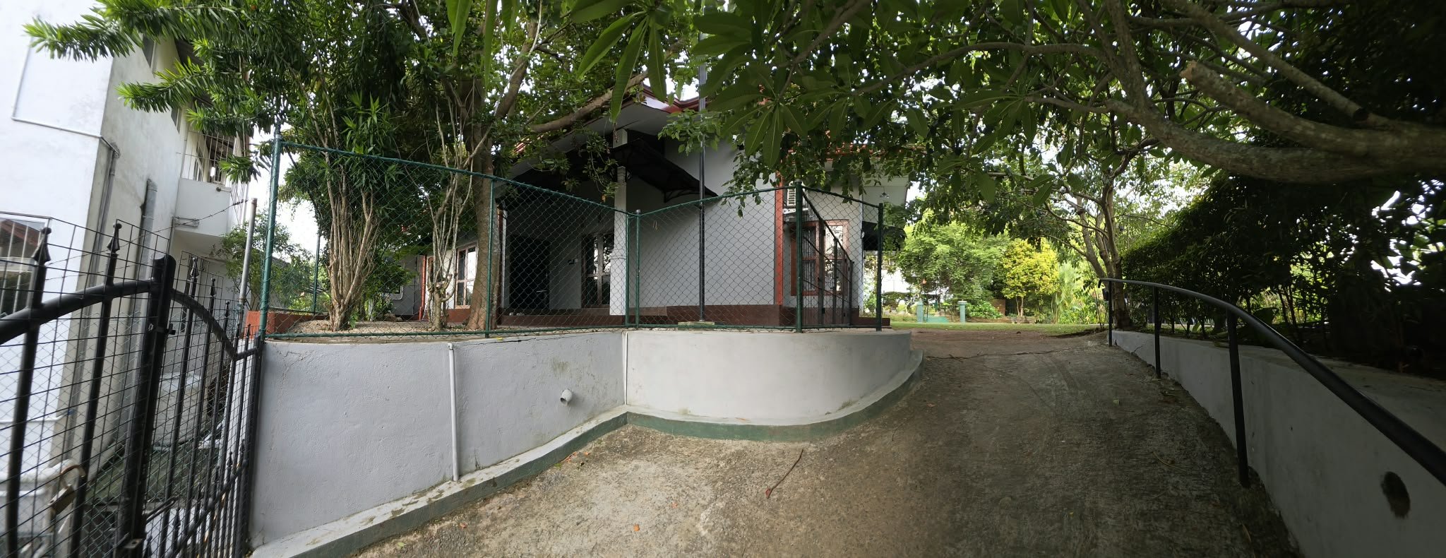A concrete pathway curves around a small, fenced outdoor area with trees and greenery, leading to a building with multiple floors and large windows, surrounded by lush trees and plants.