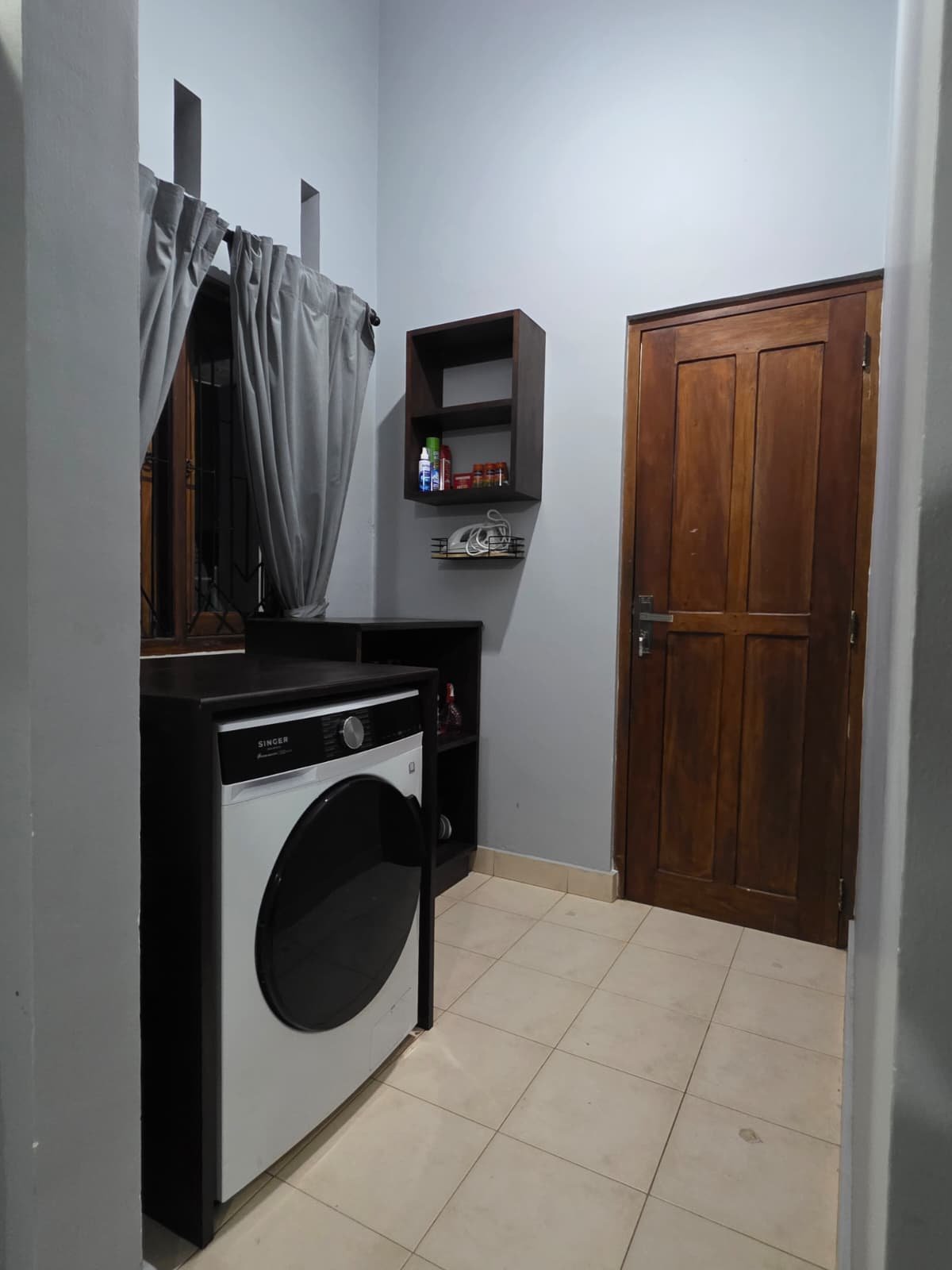A laundry room with a front-loading washing machine in black and white under a black countertop. The room has a window with gray curtains, a wooden door, and a wall-mounted shelf with laundry supplies.