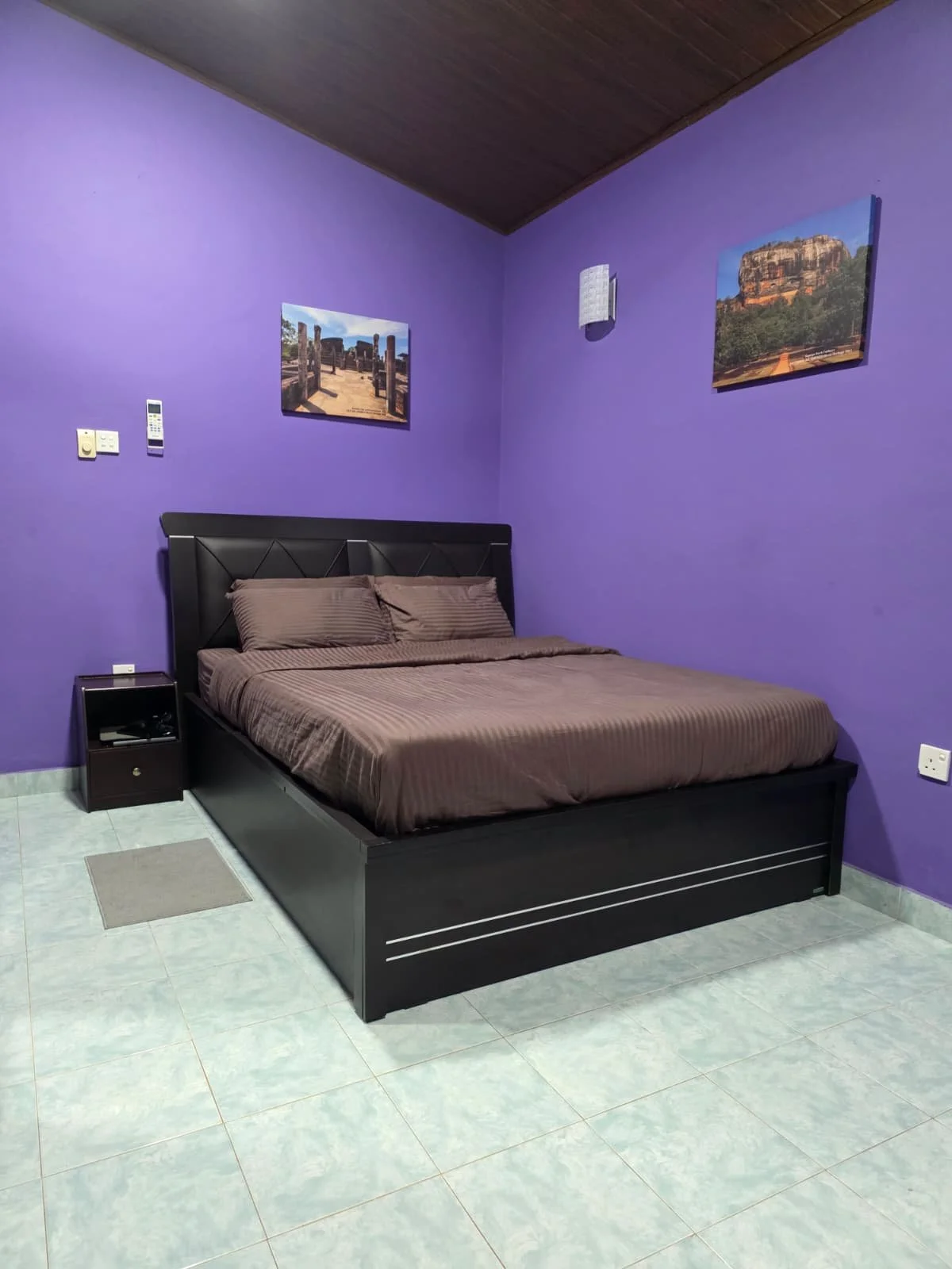 A bedroom with purple walls, a black bed with brown striped bedding, two matching pillows, a black nightstand, and three pictures of natural landscapes hanging on the walls.