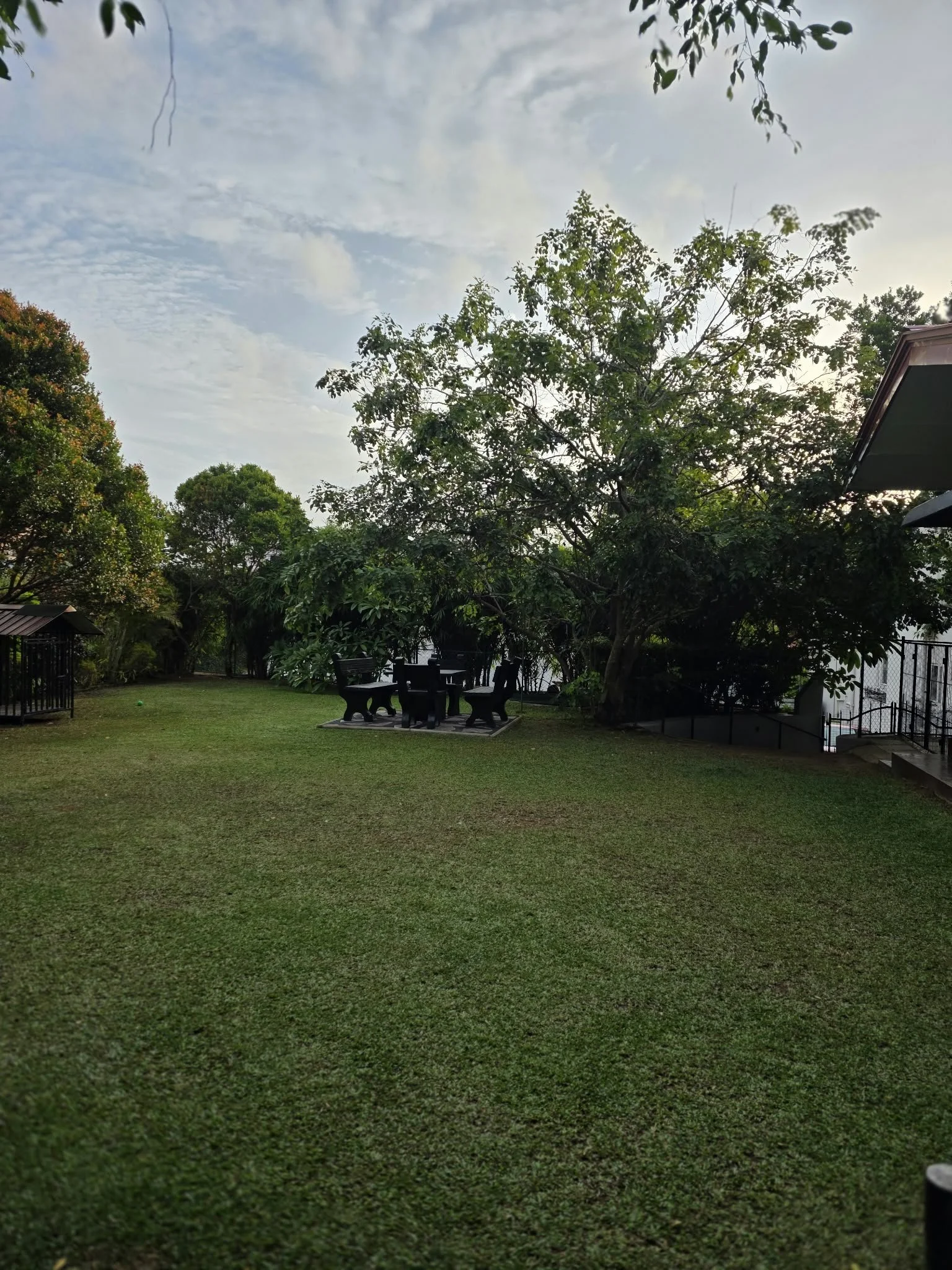 A grassy backyard area with several trees, a picnic table with benches, and some fencing in the background. The sky is partly cloudy with patches of blue.