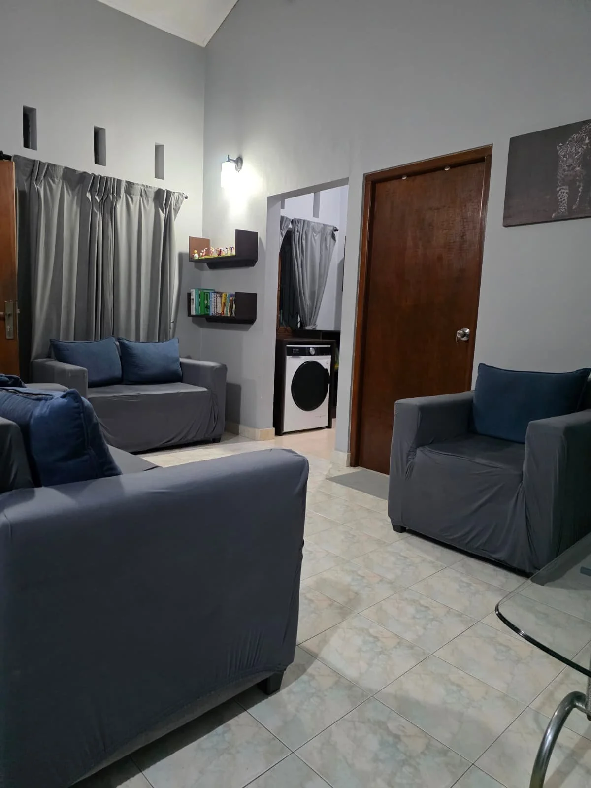 Living room with three gray sofas with blue cushions, tiled floor, a washing machine in a small open area, bookshelves, a wall-mounted light, a painting, and gray curtains on the window.