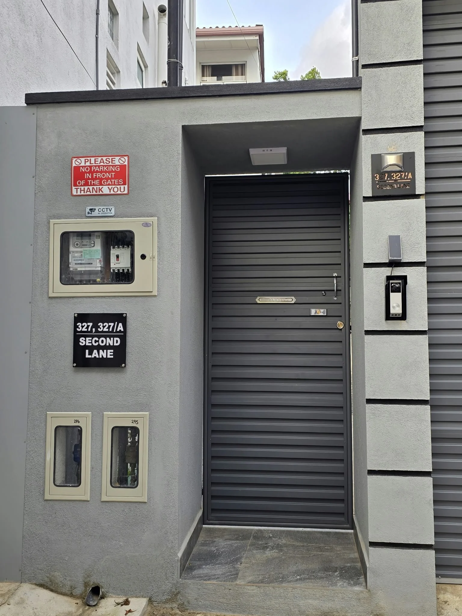 A modern building entrance with a gray metal rolling door, a black sign with white text indicating address '327, 327/A Second Lane', a gray wall with two rectangular utility boxes, a security camera sign, and a red and white no parking sign. There is