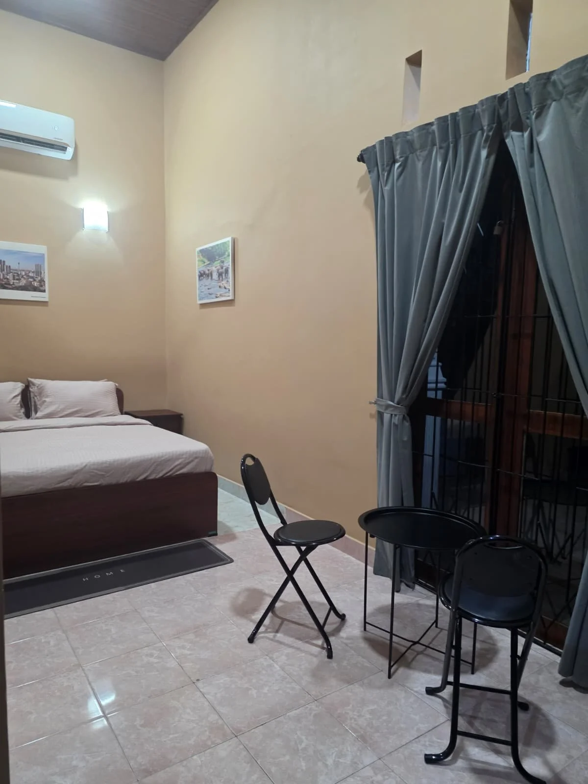 A bedroom with beige walls, a bed with white sheets, a wooden bed frame, and artwork hanging on the wall. There is a sliding glass door with gray curtains, a small round table with two black chairs, and an air conditioning unit mounted high on the wa