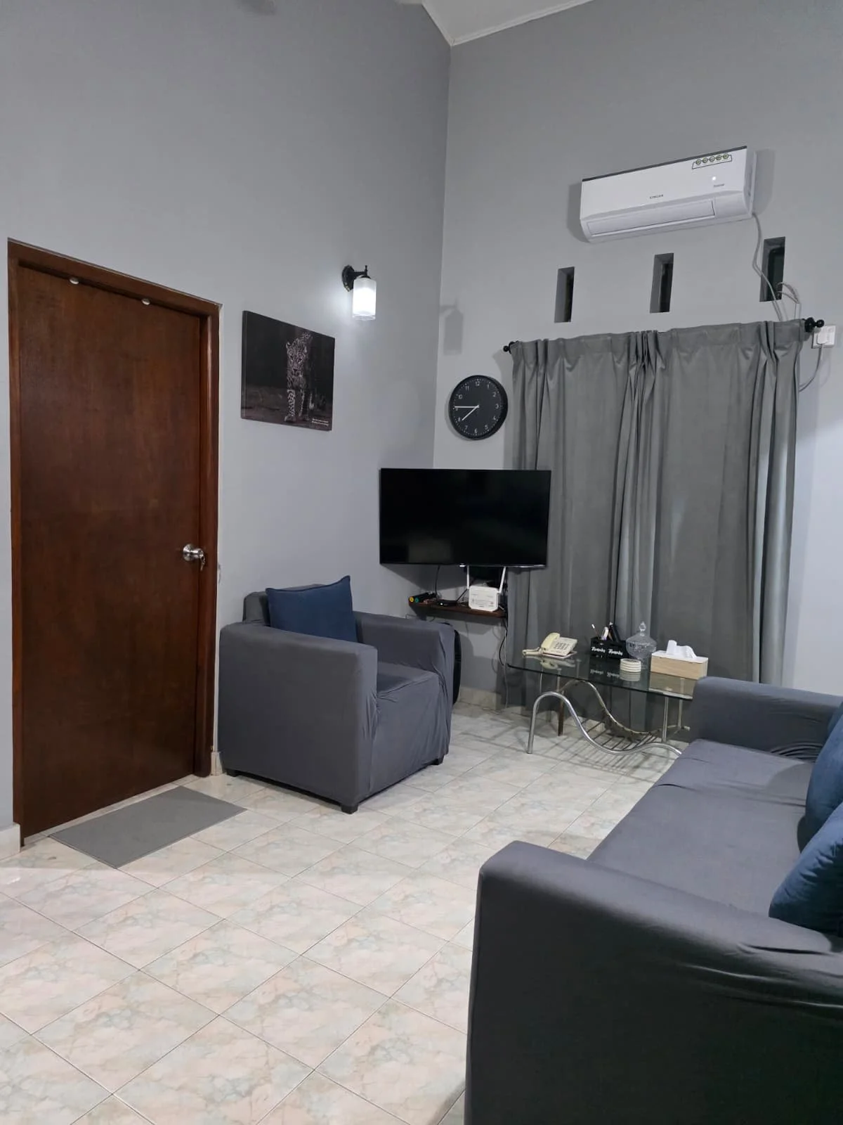A cozy living room with gray walls and tile flooring, featuring a wooden door, a wall-mounted light, a framed photo, a black wall clock, a flat-screen TV on a small wall-mounted shelf, two gray armchairs with navy pillows, a glass coffee table with i