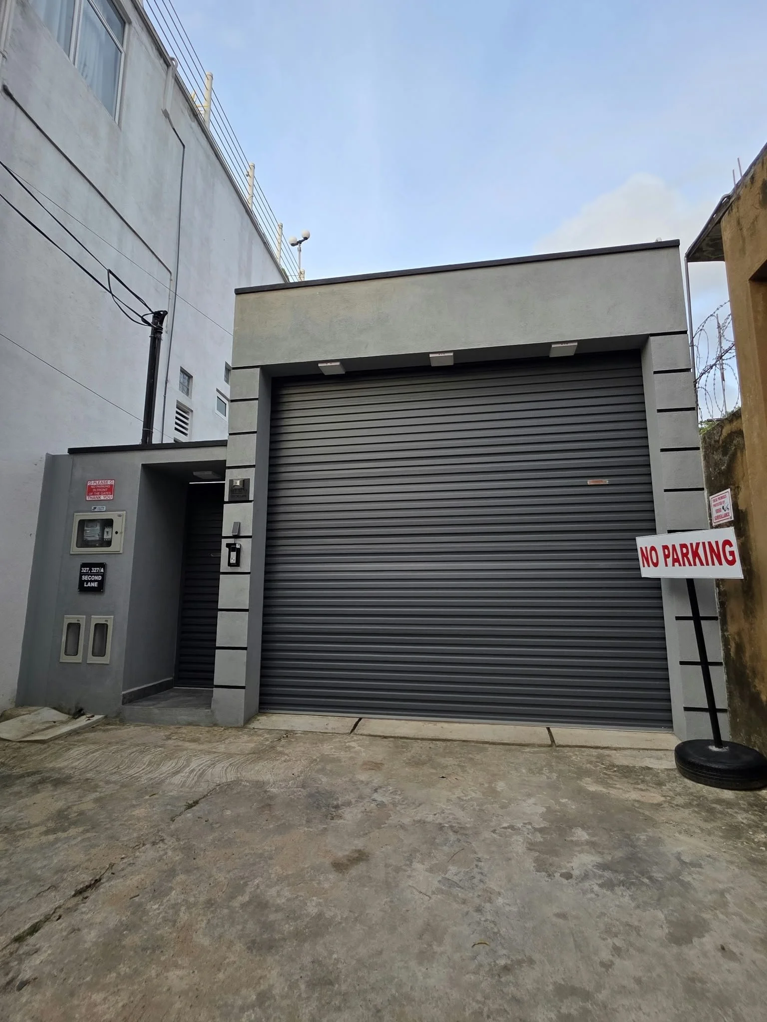 A gray building with a closed metal garage door, a small window, and electric meters outside. There is a 'No Parking' sign and a narrow pathway in front. The concrete ground is uneven and slightly worn.
