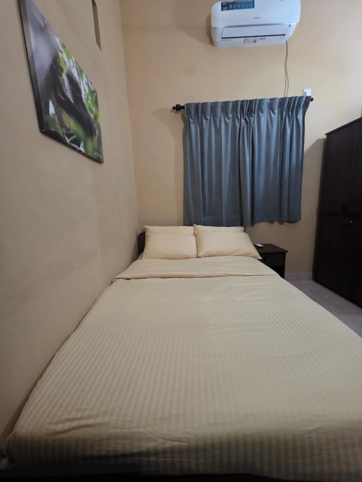 A bedroom with a bed, cream-colored bedspread, two pillows, a small black nightstand, a dark wooden wardrobe, a window with blue curtains, an air conditioning unit, and a wall-mounted painting of a landscape.
