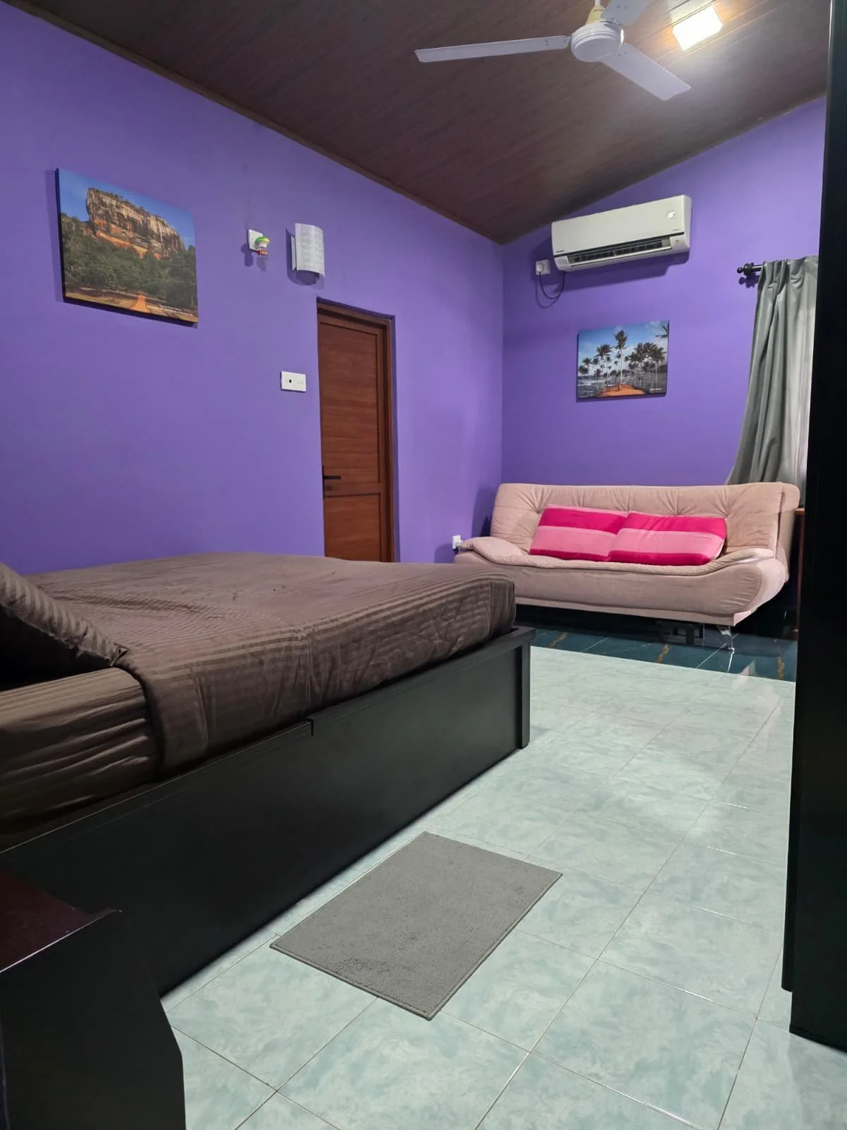 A living room with purple walls, a brown bed, a beige sofa with pink cushions, a wooden door, wall art, an air conditioning unit, a ceiling fan, and curtains.