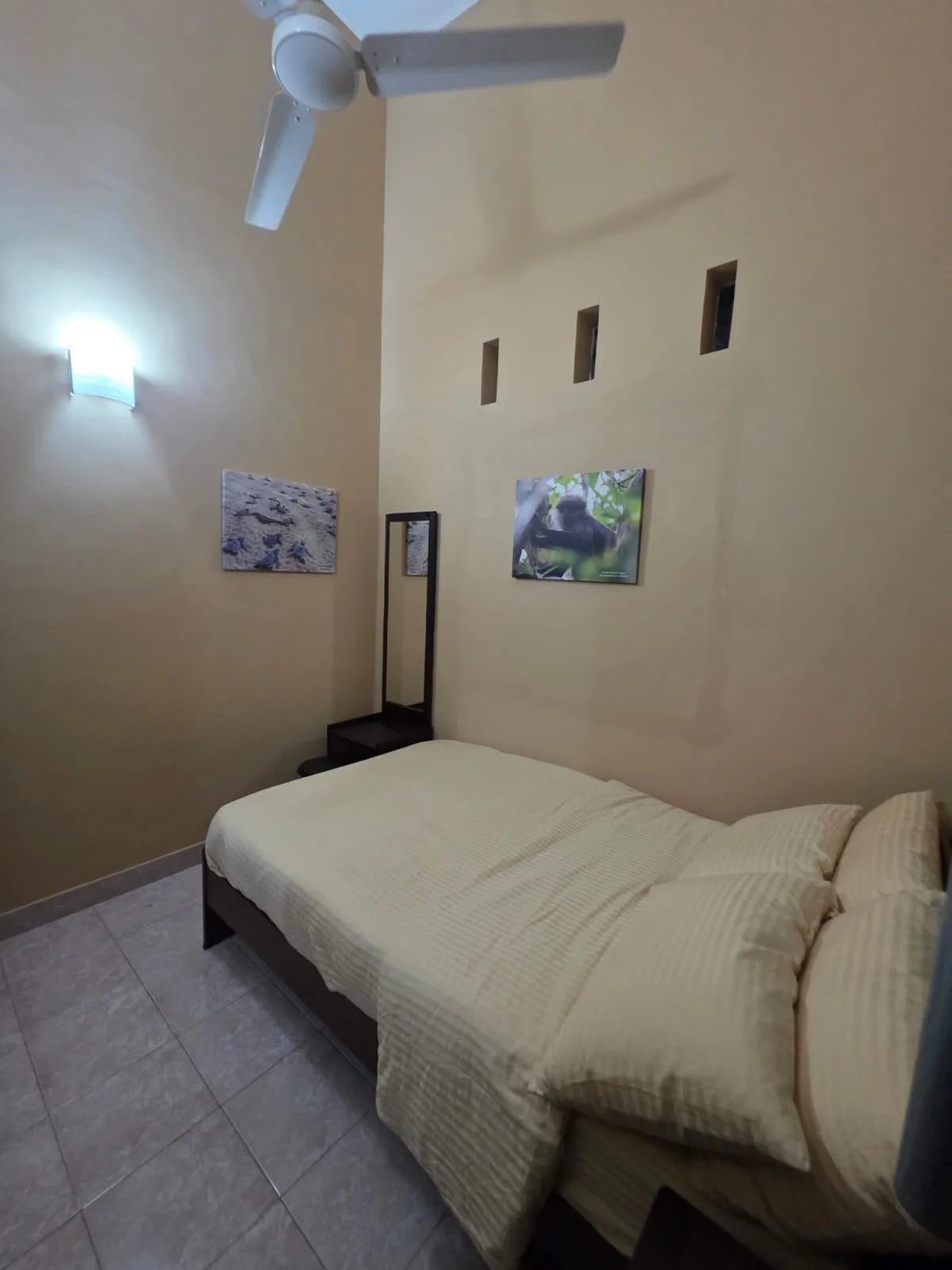 A modest bedroom with a bed covered in a beige striped blanket, a small black nightstand with a mirror, yellow walls decorated with framed pictures, a wall-mounted light, a ceiling fan, and three small square wall openings.