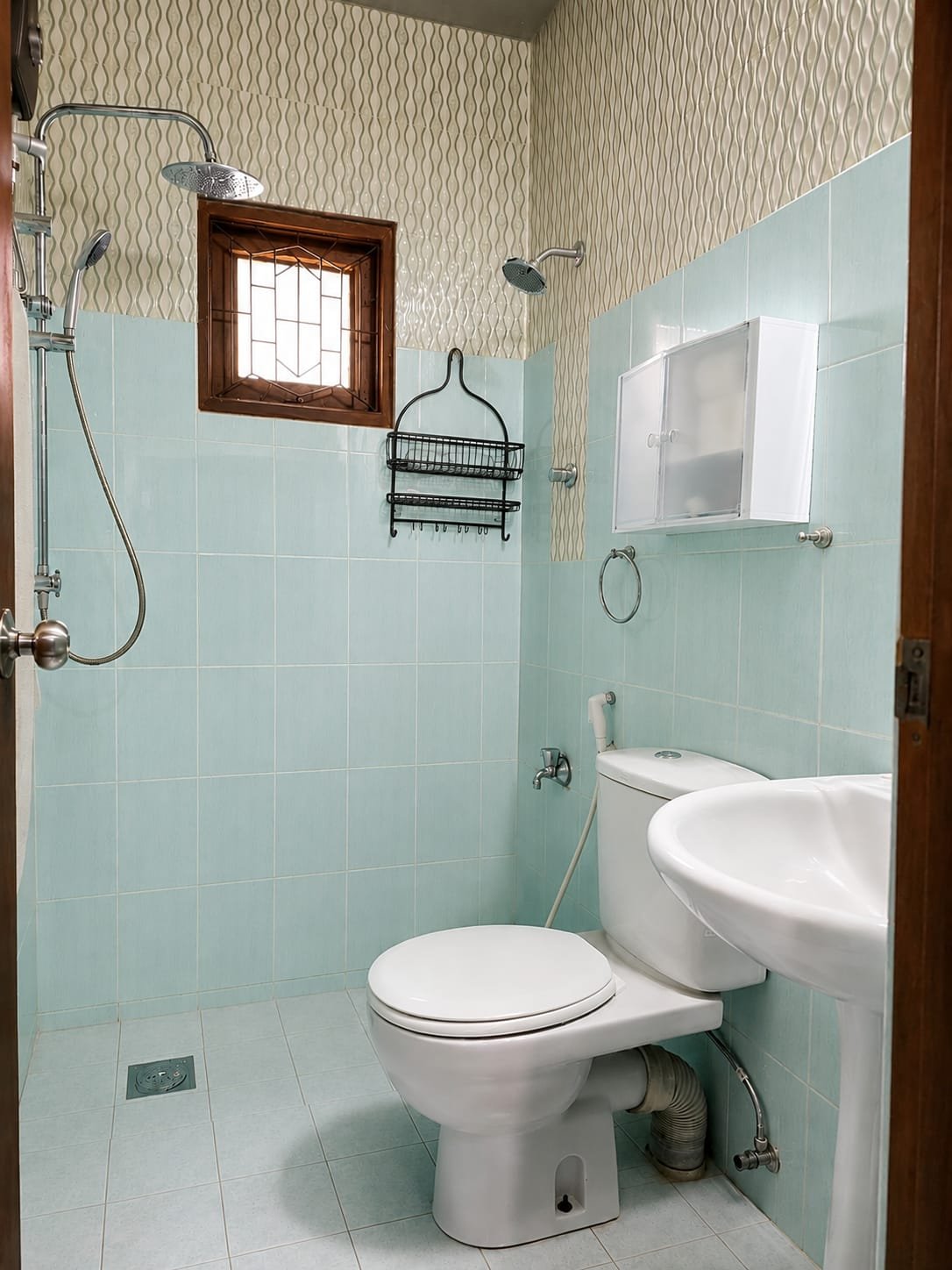 Small bathroom with light blue tiled walls, a toilet, a small white sink, a wall-mounted white cabinet, a towel ring, a window with wooden frame, and two shower heads with a shower caddy.
