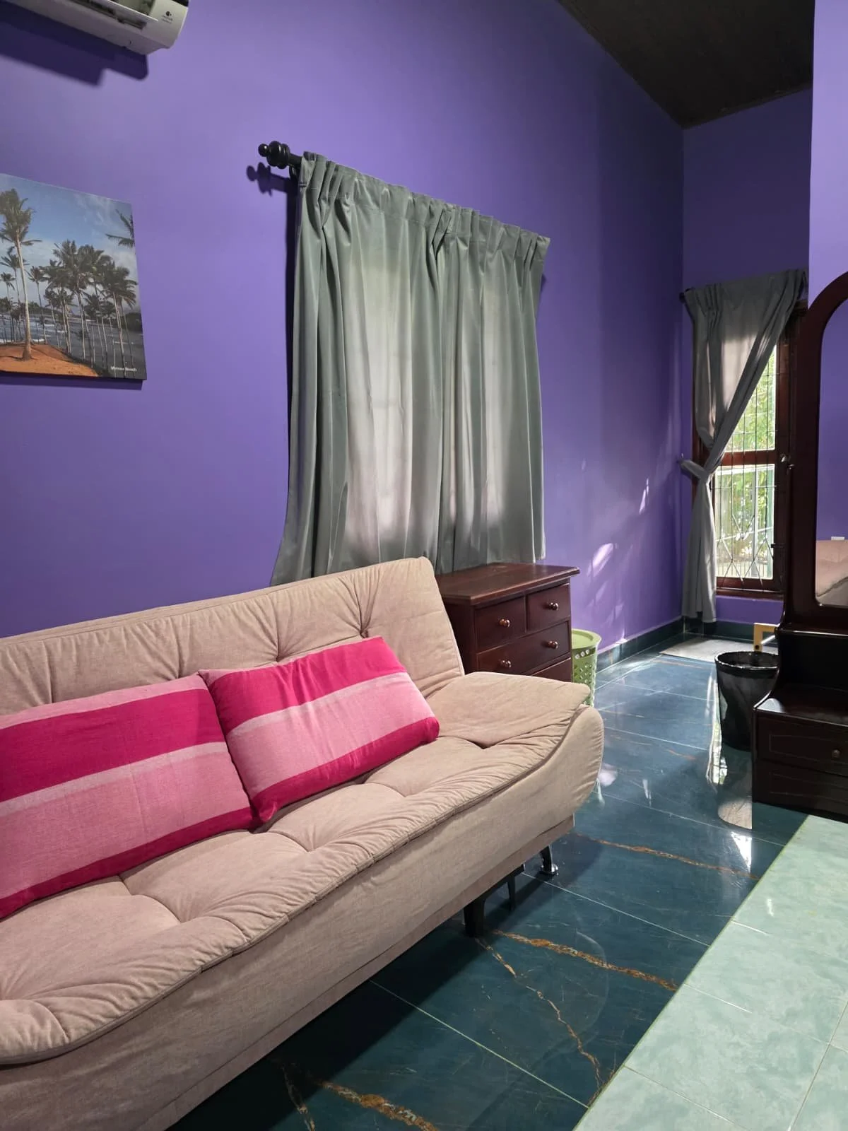 A living room with purple walls, beige sofa with pink and red striped pillows, dark wood furniture, green curtains, and a window with bars letting in natural light.