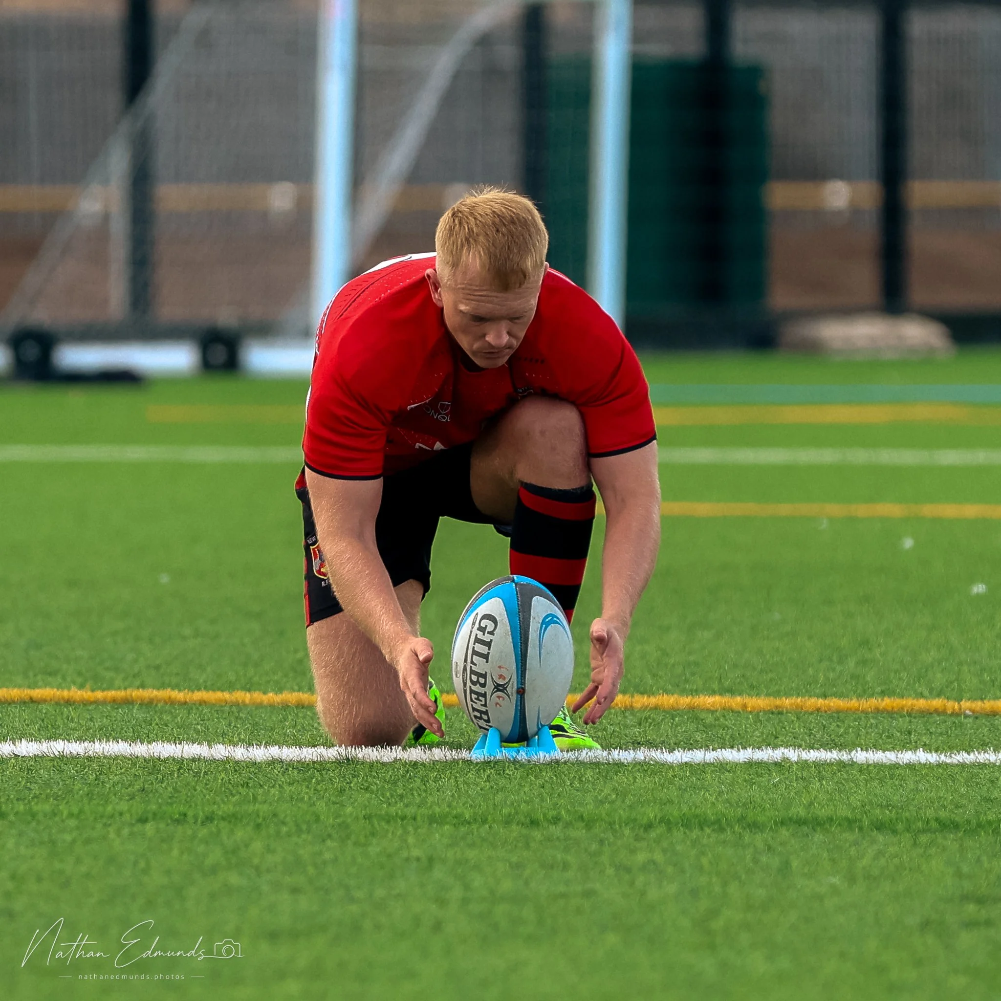 A rugby player in a red jersey and black shorts is on his knees on a green field, holding a rugby ball while a teammate supports him from behind. An outdoor sports stadium with a fencing and seating area is visible in the background.
