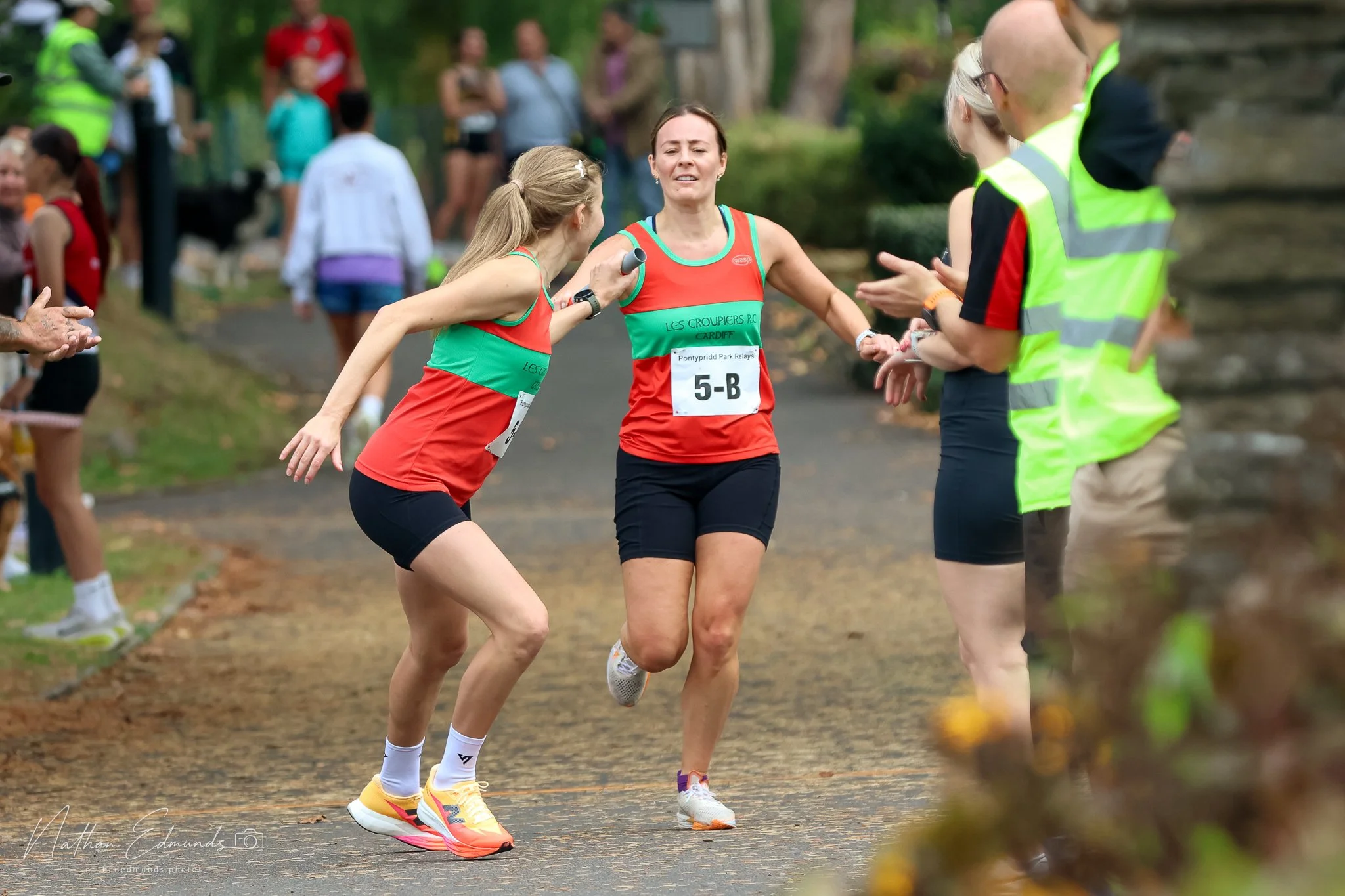 Two female runners crossing a finish line at a race, with one woman in red and green athletic wear and the other in red and black, while a woman in a yellow vest and glasses hands them medals, in a park with a dirt path and spectators in the background.