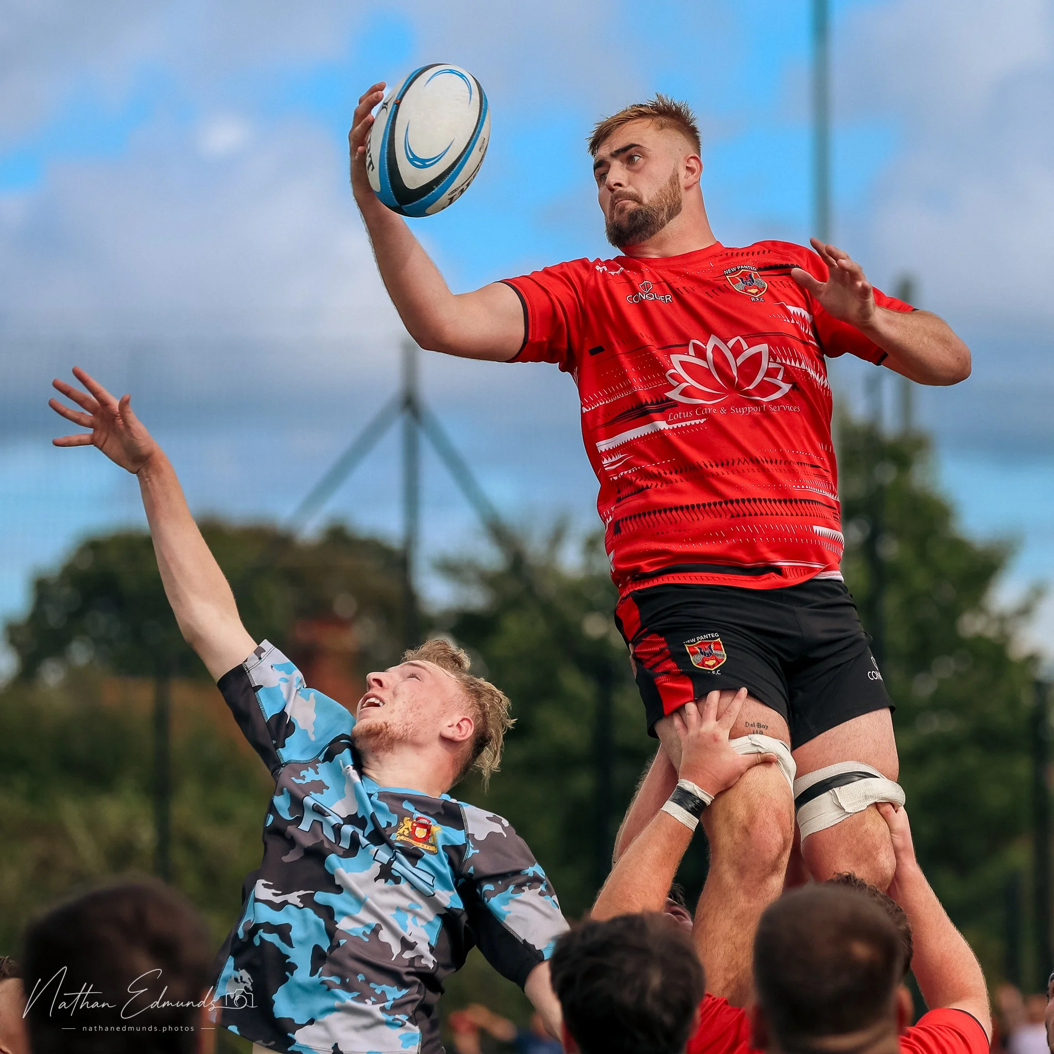 A rugby player in a red jersey is leaping into the air and holding a rugby ball, while a player in a camouflage jersey is reaching up with one arm. The scene appears to be outdoors with a mostly cloudy sky and trees in the background.