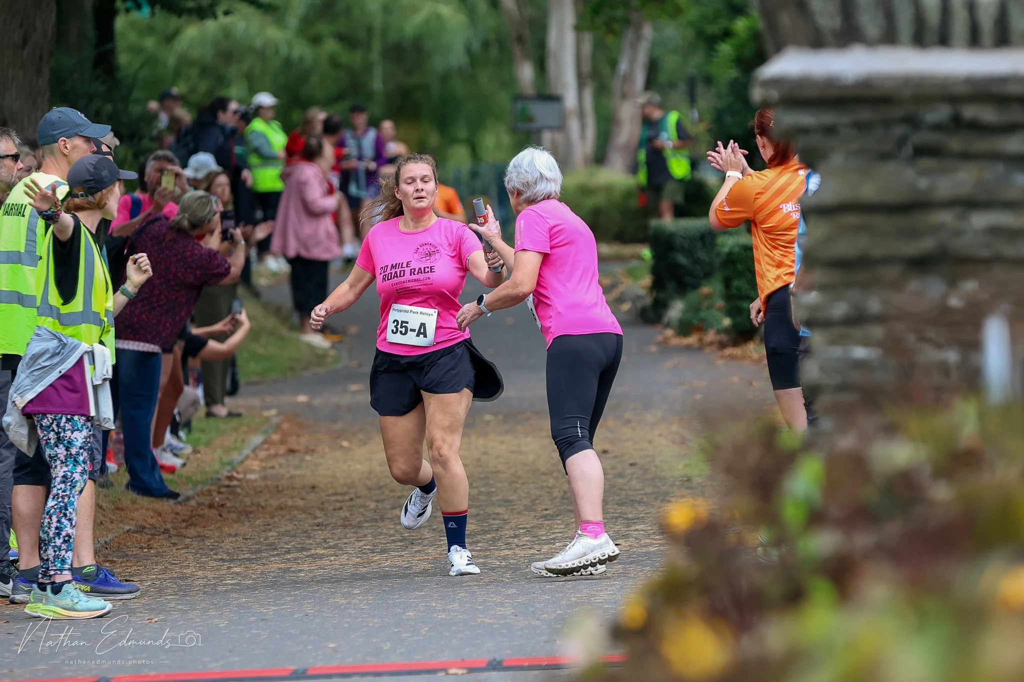 A woman in a pink shirt and black shorts running in a race while an older woman in a matching pink shirt hands her a water bottle. Multiple spectators and supporters line the street, some taking photos. The scene is outdoors with trees surrounding the pathway.