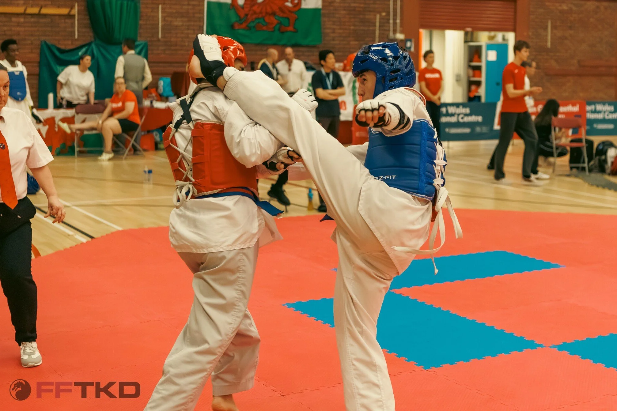 Two martial artists in middle of a sparring match in a gymnasium, wearing white uniforms, protective helmets, and gloves, with spectators and officials in the background.