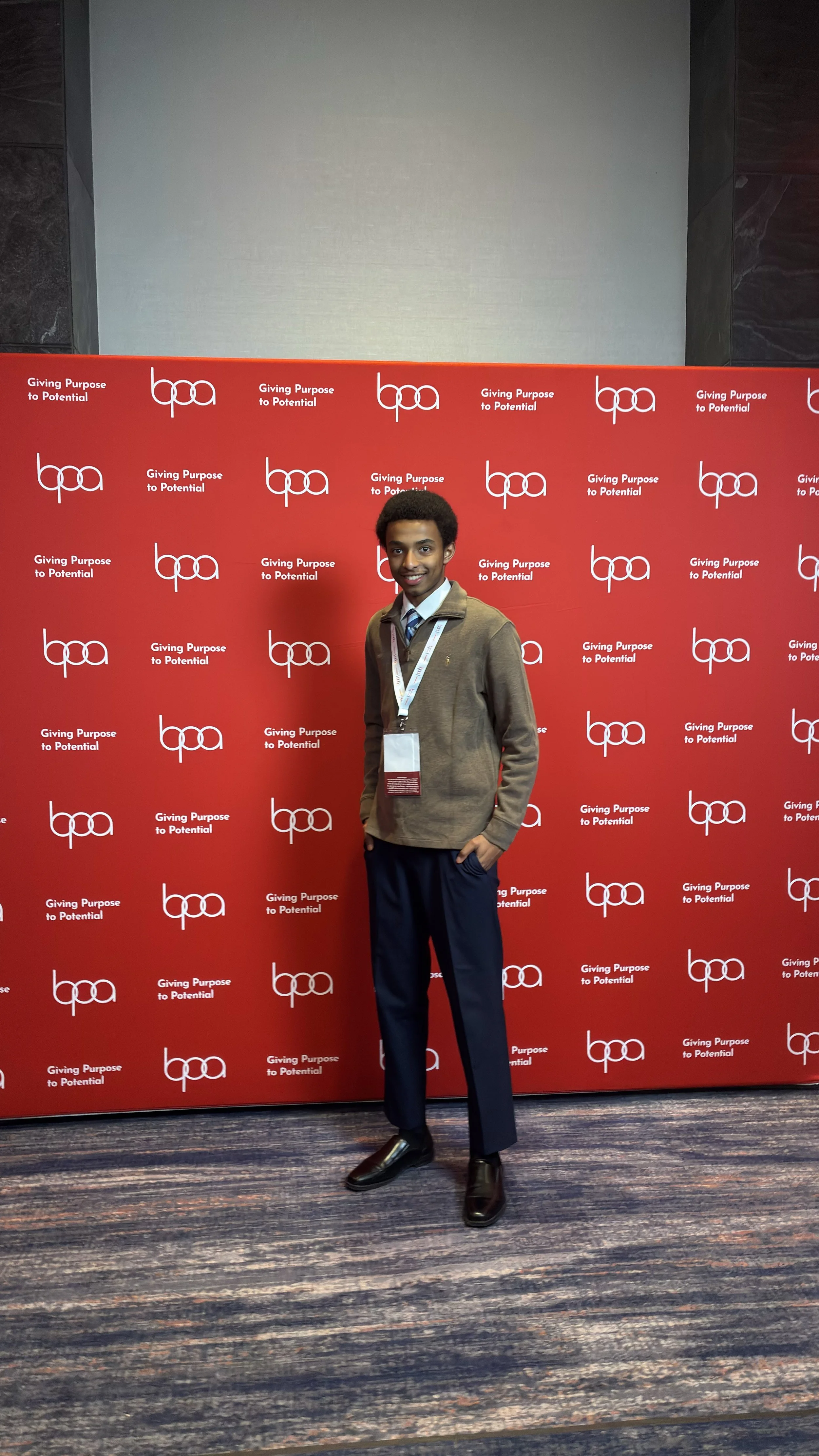 A young man standing in front of a red backdrop with white logos and text reading "Giving Purpose to Potential." He is wearing a beige jacket over a collared shirt and tie, black dress pants, and black shoes, and has a white lanyard around his neck.