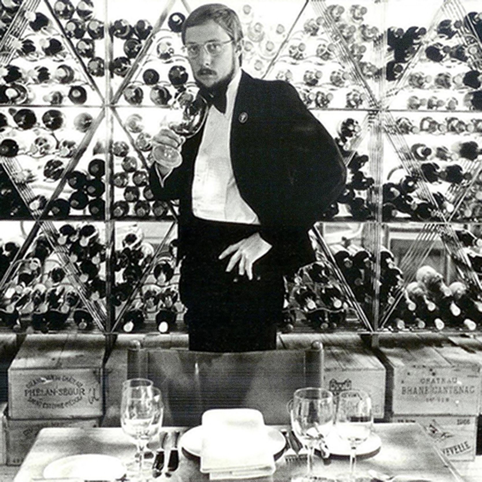 A man in a tuxedo and glasses holding a wine glass, standing in front of shelves filled with wine bottles at a wine tasting event.