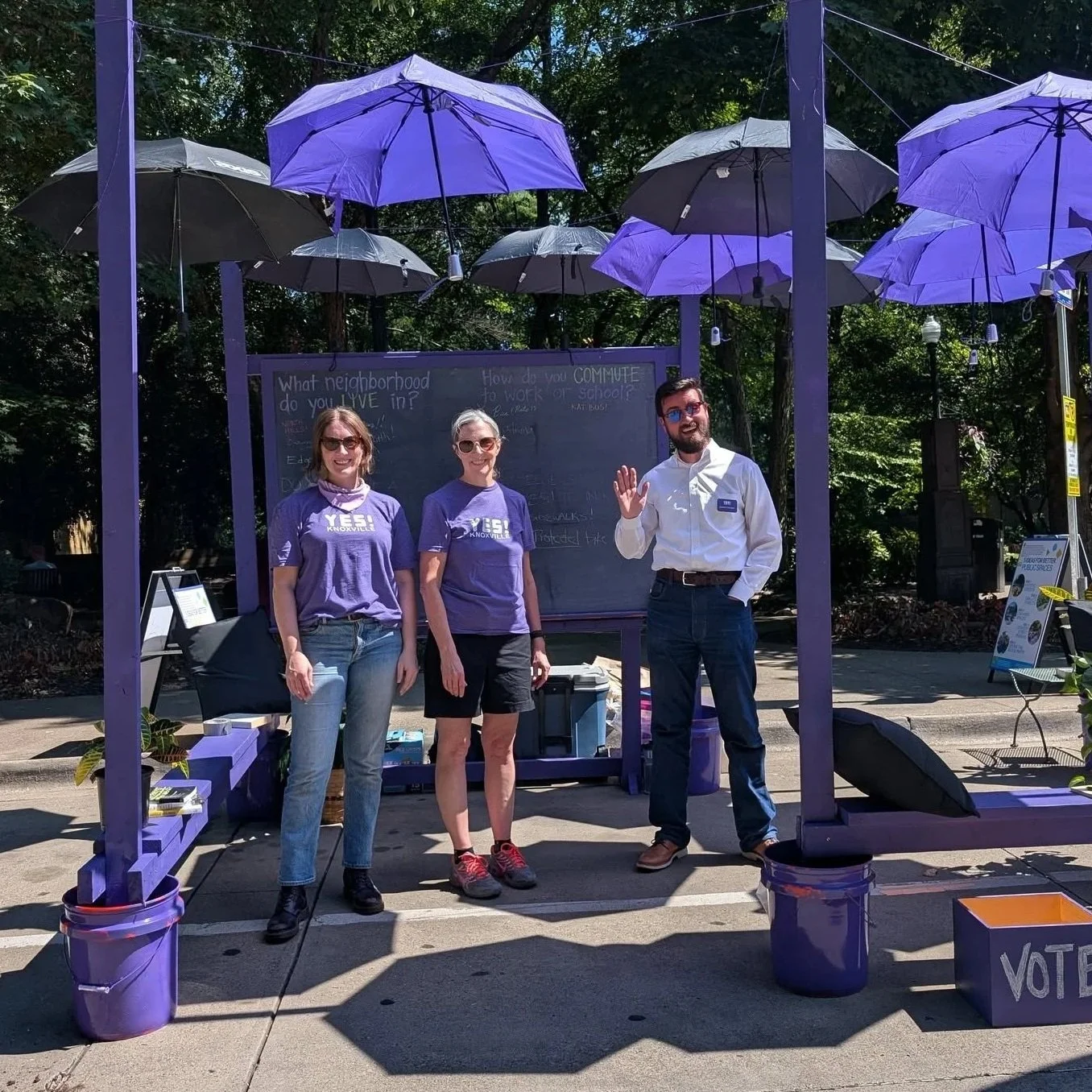 Three people standing inside a purple booth with umbrellas overhead at a Parking Day event in Downtown Knoxville. Two women in purple shirts and a man in a white shirt are smiling, with the man waving.