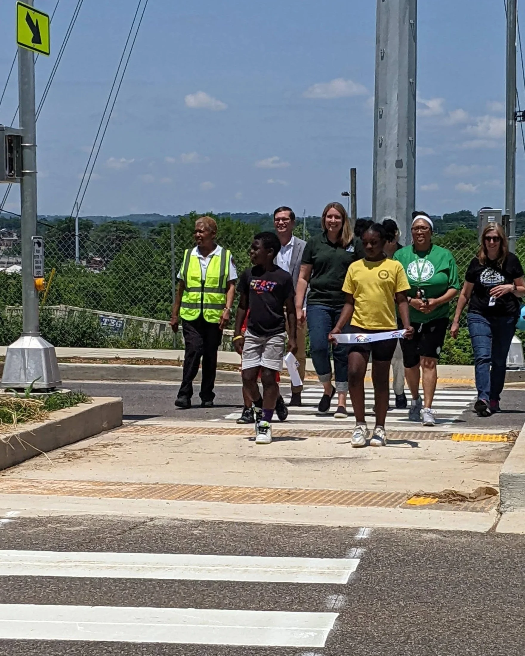 Group of people walking across a pedestrian crosswalk in East Knoxville, some smiling, a mix of genders and ages, with trees and a sunny blue sky in the background.
