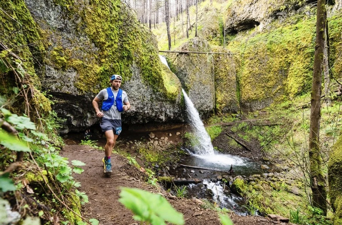 A man runs in the forest. There is a waterfall behind him.