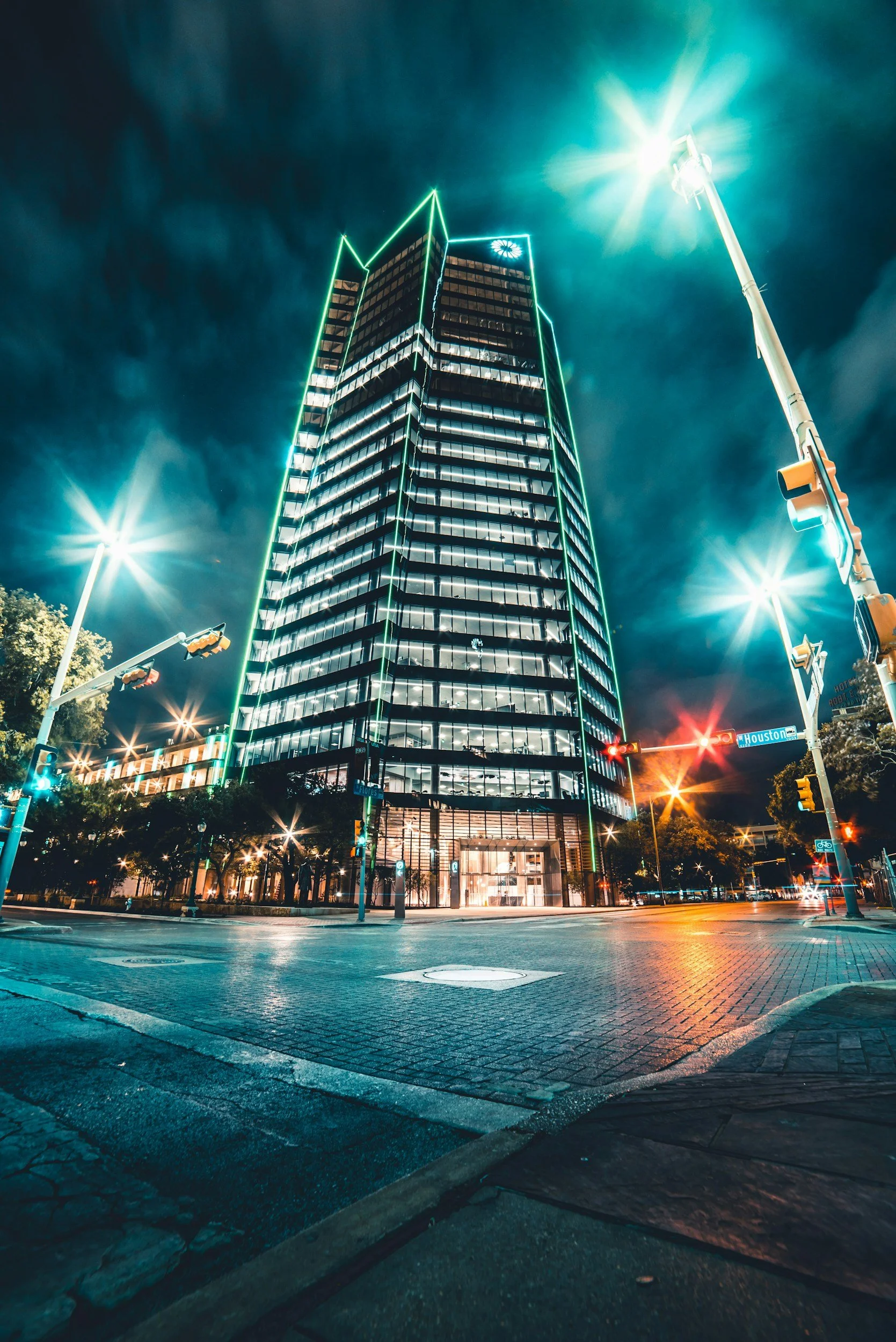 Night view of a tall modern office building with illuminated windows and green neon outlines, located at an intersection with traffic lights and streetlights, reflecting lights onto the wet pavement.