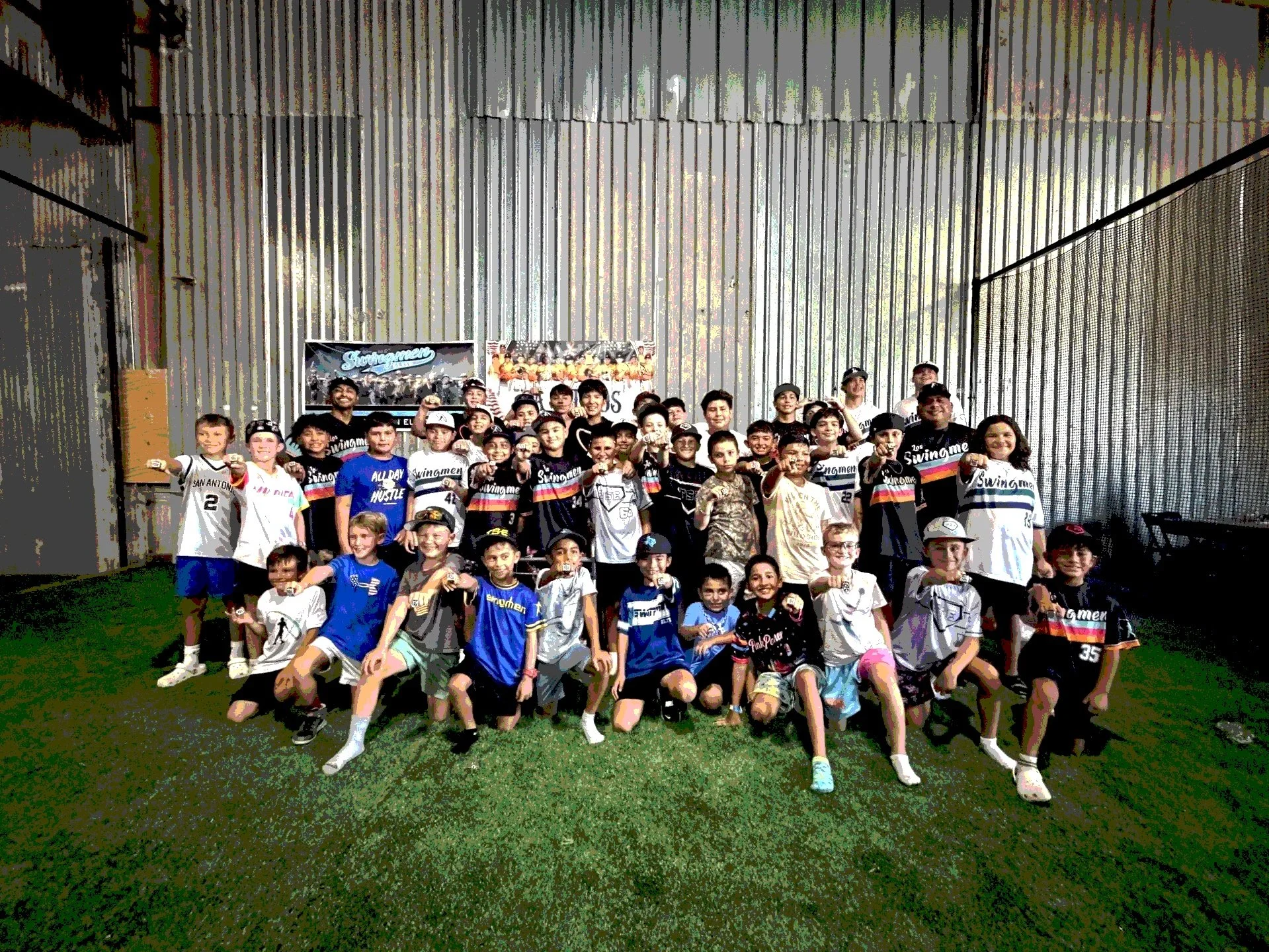 A large group of children and a few adults posing for a photo on an indoor field with green artificial grass. Many children are wearing sports jerseys and hats, some are holding medals. The background shows a metal wall and a digital screen with the word 'Swingen' displayed.