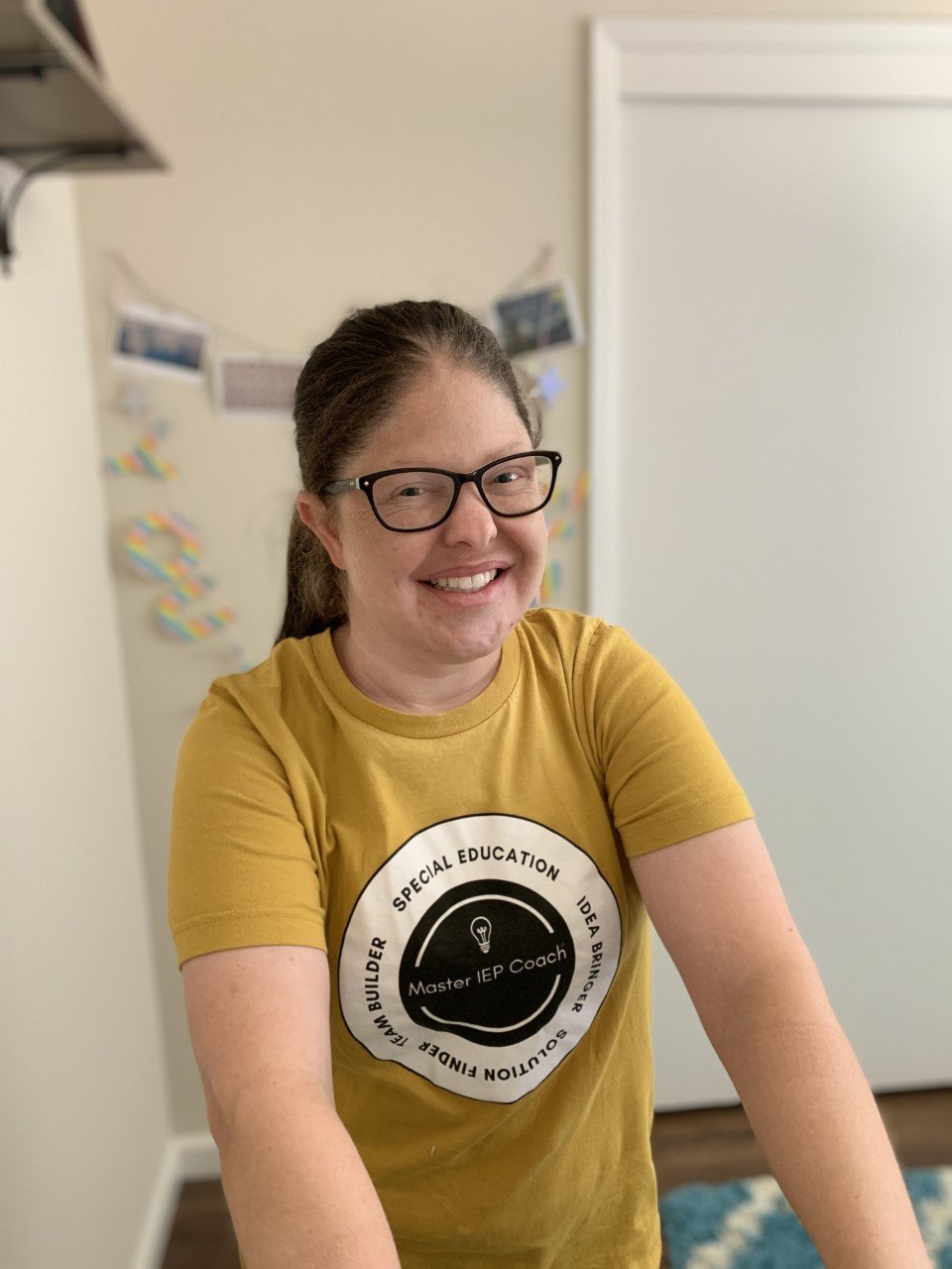 A woman with glasses wearing a yellow shirt, smiling, with a blurred background of a room.
