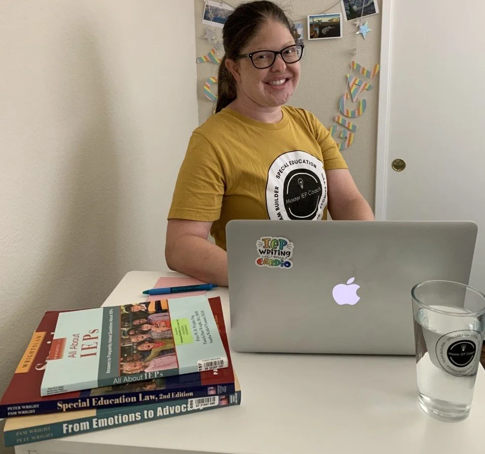 A woman wearing glasses and a yellow T-shirt sitting at a table with books, a MacBook laptop, a pen, and a glass of water with a sticker, smiling at the camera.
