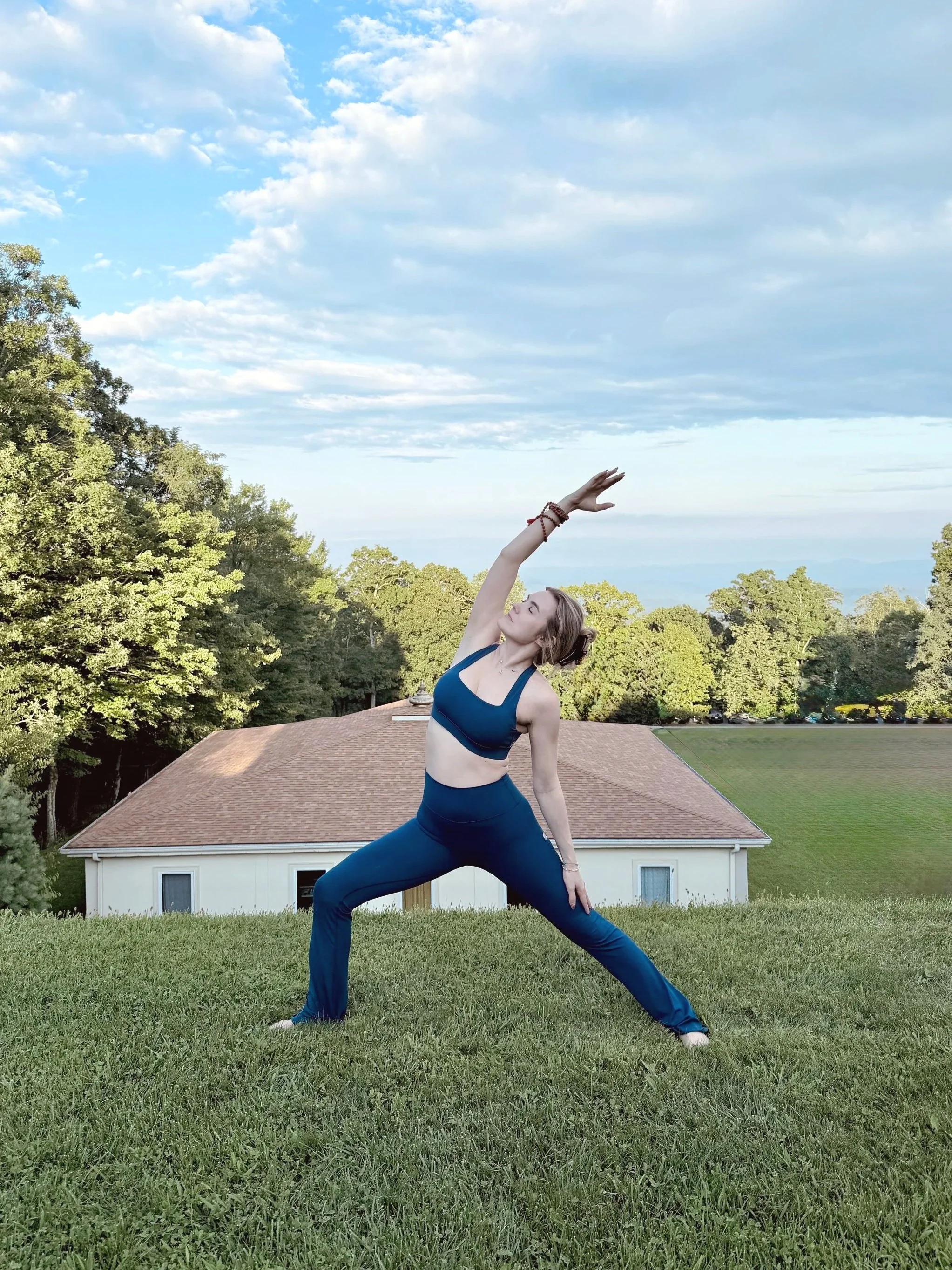 A woman practicing yoga outdoors on a grassy lawn, wearing a blue sports bra and matching leggings, with a house and trees in the background under a partly cloudy sky.