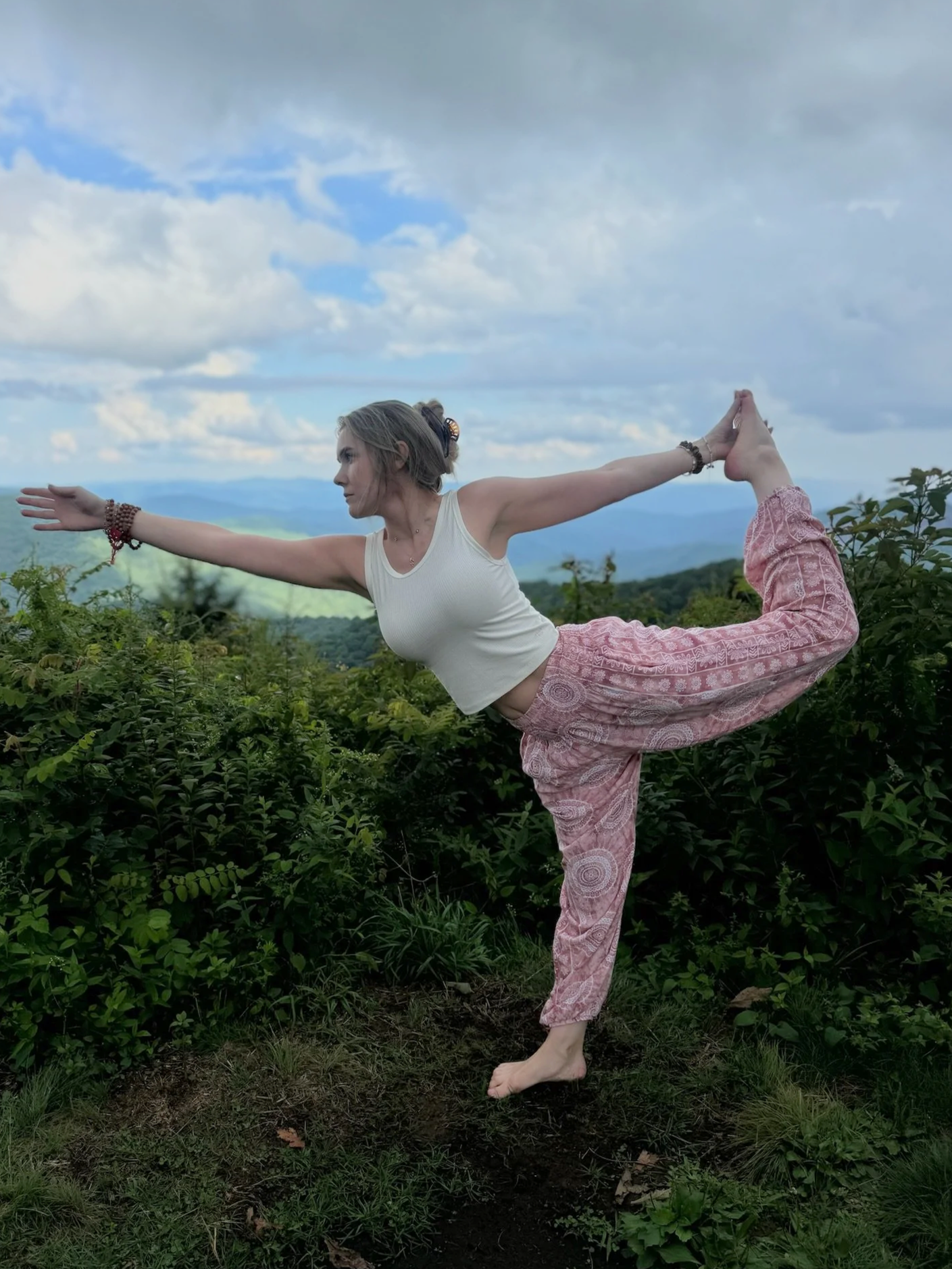 A woman practicing yoga outdoors on a hillside with mountains in the background, standing on one leg with her other leg bent backward and held with her hand, forming a balanced pose.