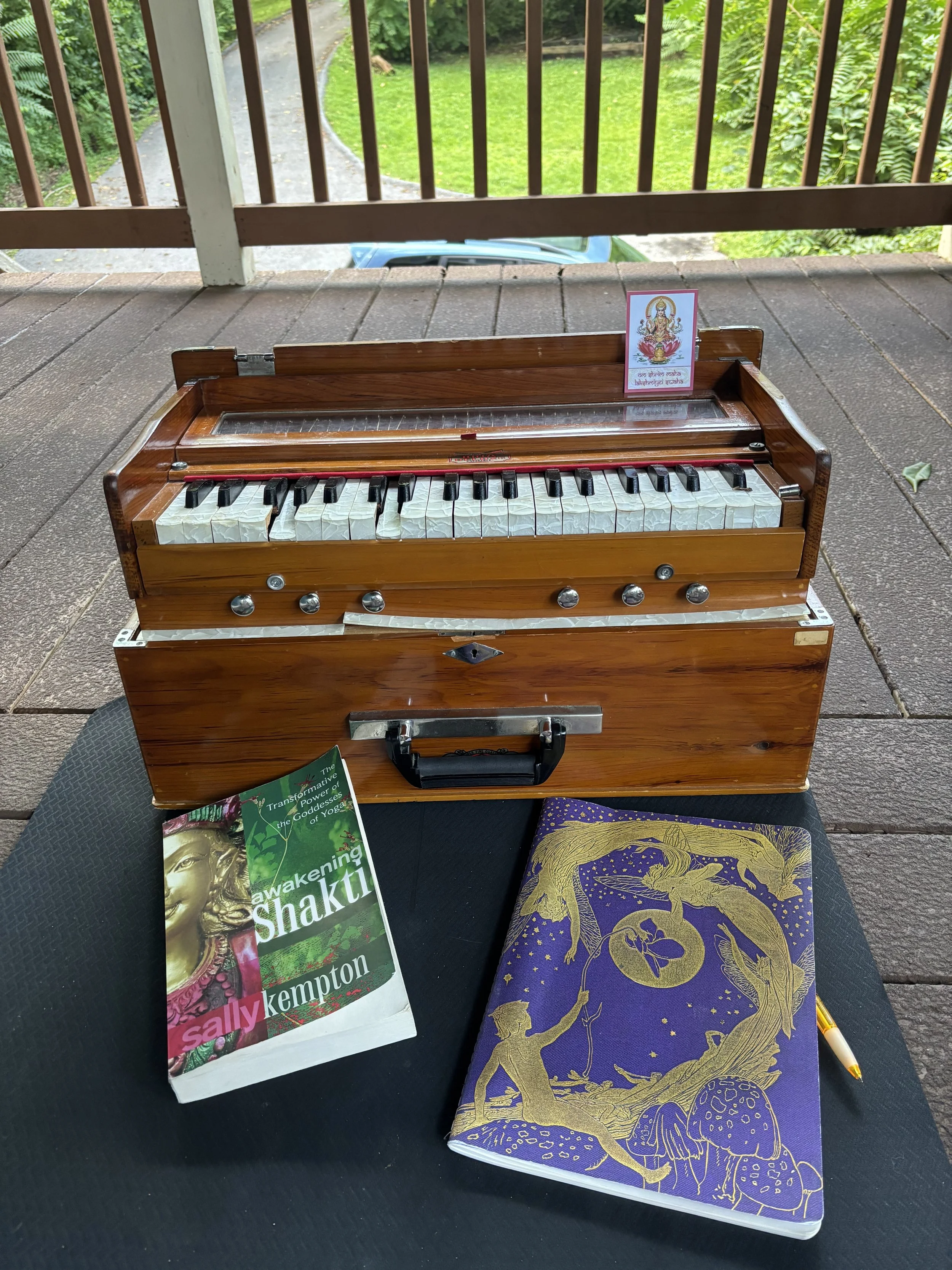A wooden toy piano with a small stand, a book titled "Awakening Shakti" by Sally Kempton, a purple notebook with gold illustrations of fairies and mushrooms, a yellow and black patterned pen, and a small card with a colorful Hindu goddess illustration, placed on a black surface outdoors with green grass and foliage in the background.