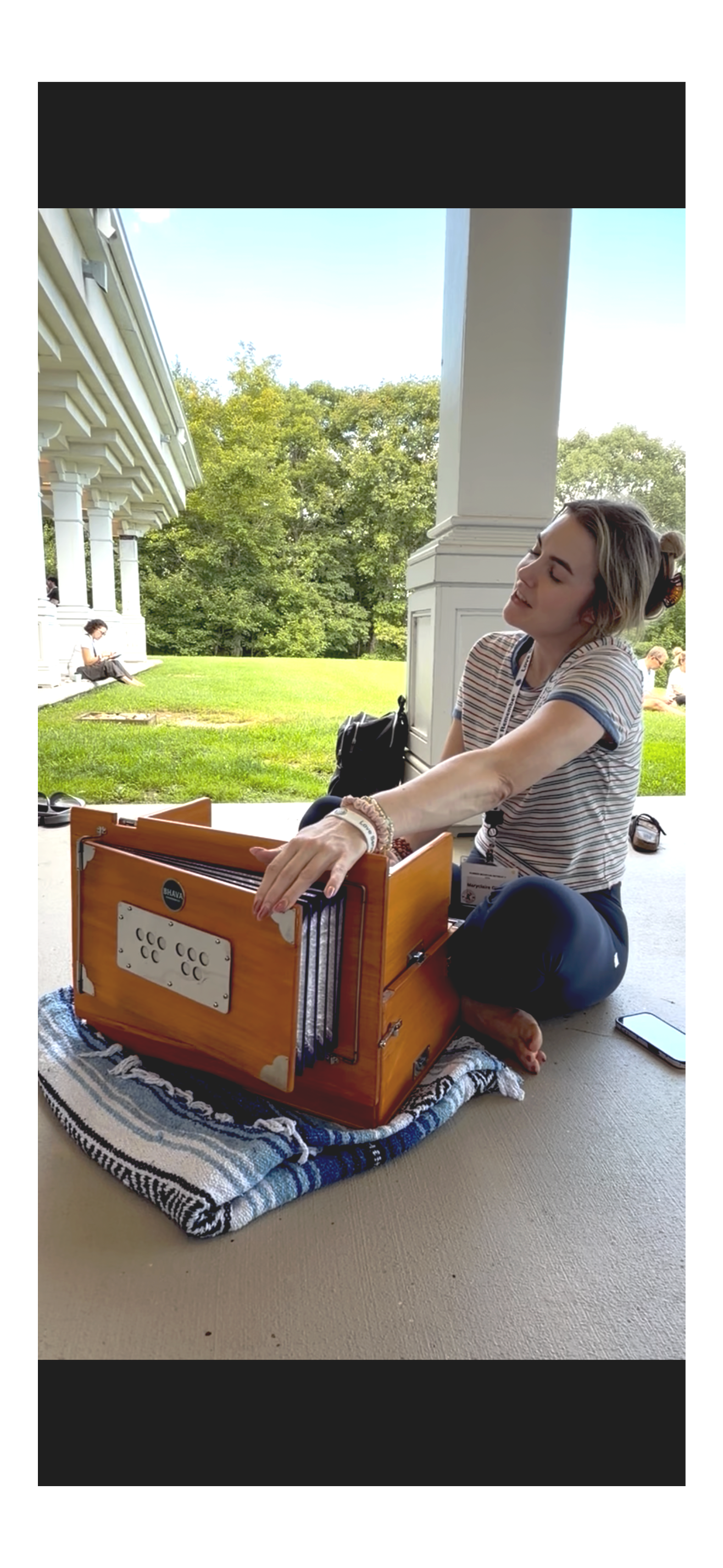 A woman playing a hammered dulcimer outdoors on a porch, sitting cross-legged on a blanket, wearing a striped shirt and wristbands. In the background, a girl is sitting on the grass reading, with trees and a Victorian-style house with columns visible.