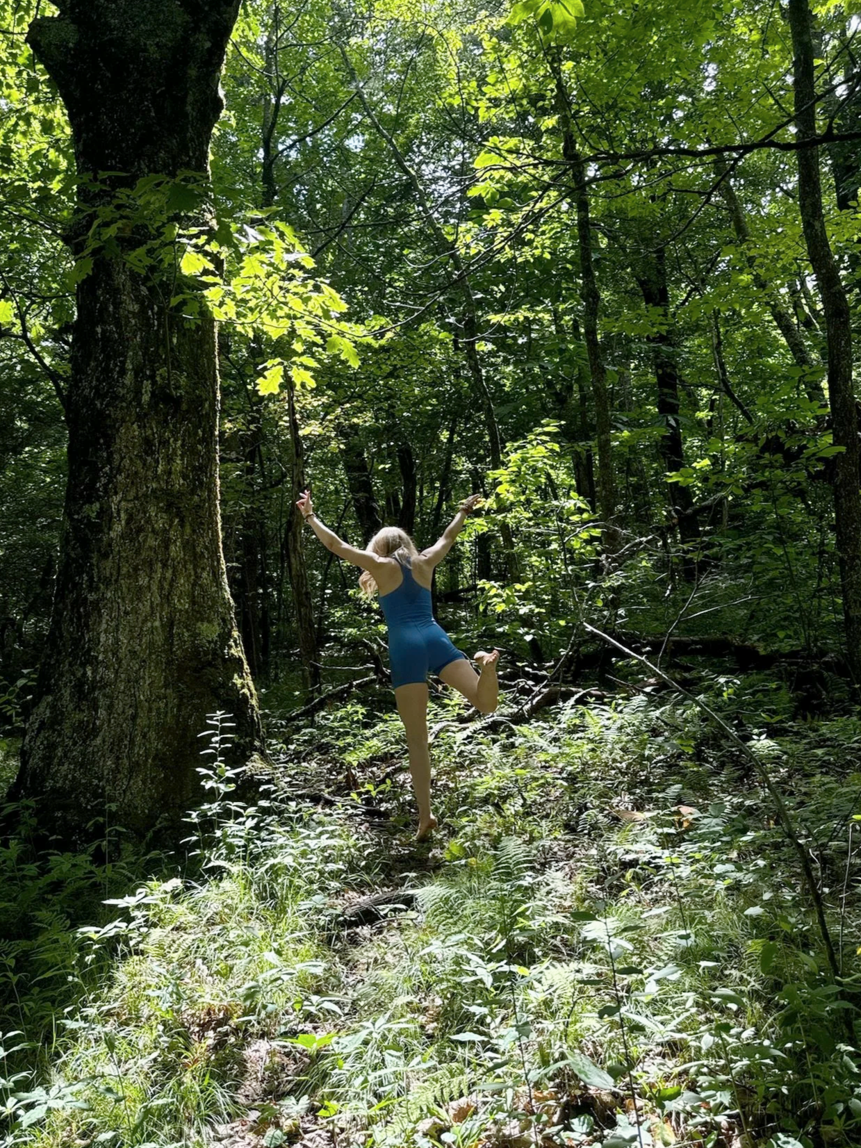 A person in a blue swimsuit is standing on one leg with arms outstretched in a lush green forest, reaching towards the sky.