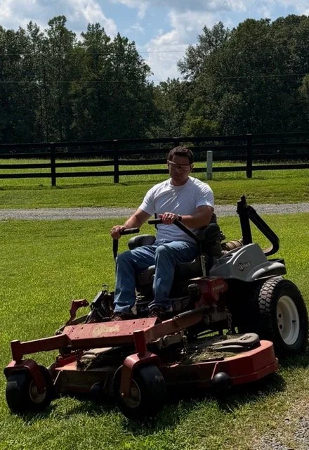 Young man sitting on a riding lawn mower on a grassy field with trees and a fence in the background.