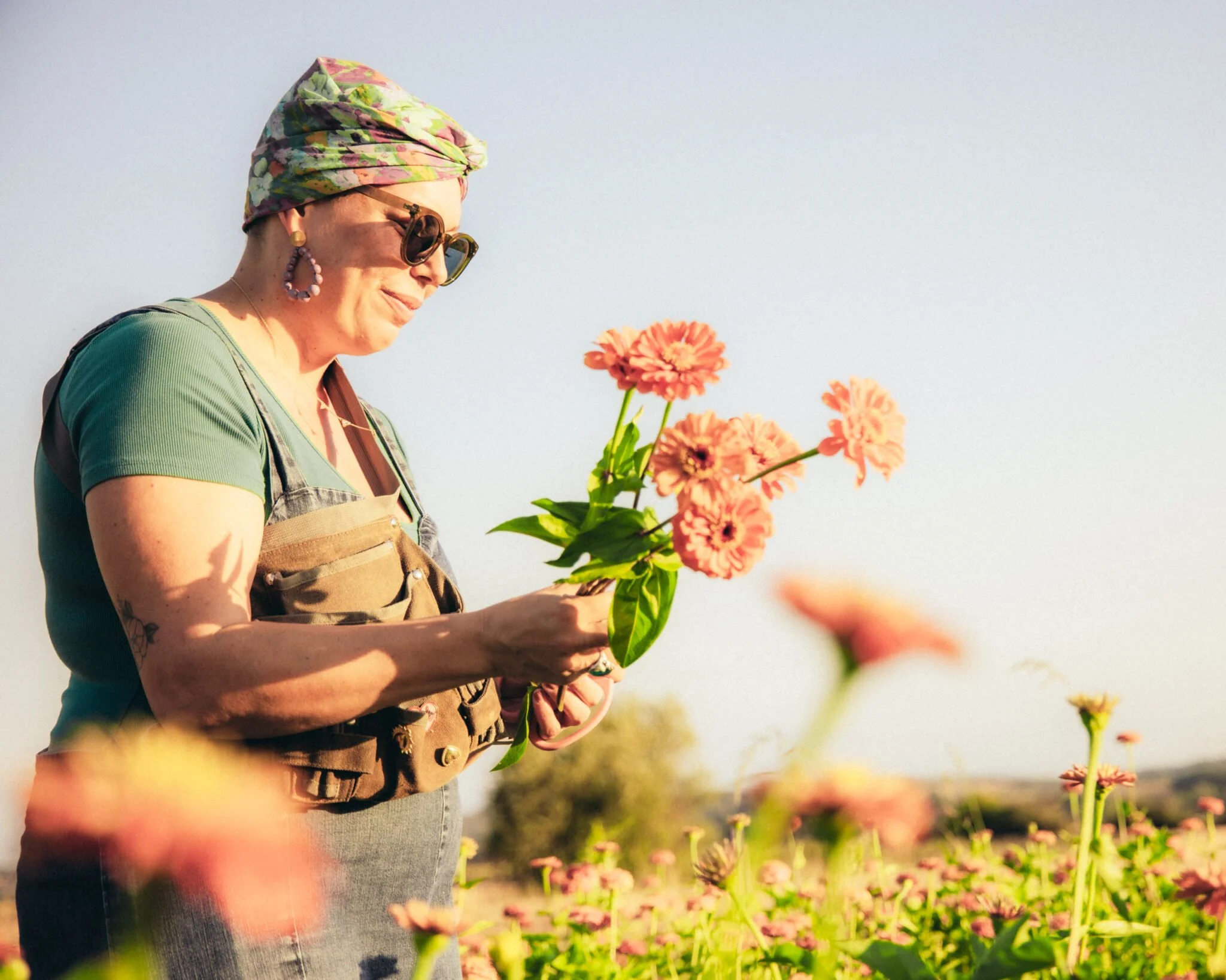 Une femme portant une coiffe colorée, des lunettes de soleil, un haut vert et un tablier, tient un bouquet de fleurs dans un champ ensoleillé.