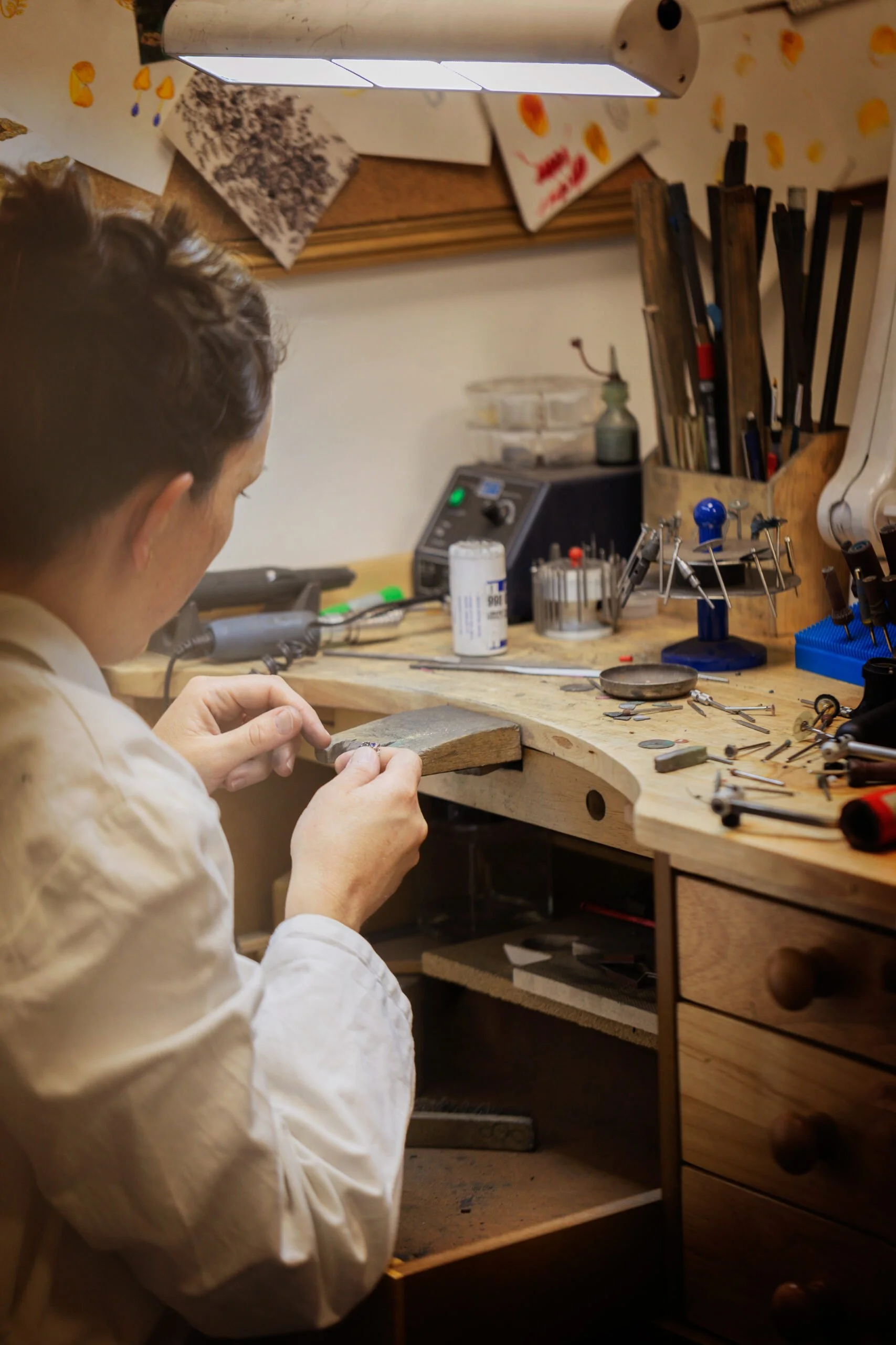 Une personne travaille dans un atelier de bijouterie, manipulant un objet avec ses mains, entourée d'outils et de matériel de travail sur un plan de travail en bois.